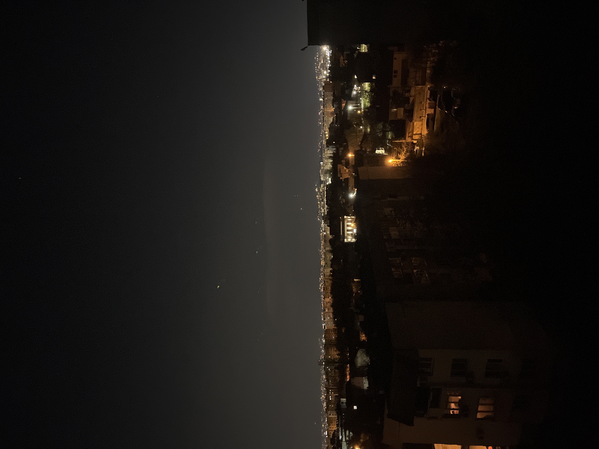 This is an outdoor, high-angle photograph taken at night. The view overlooks a city, presumed to be Sofia, Bulgaria, as indicated by the request, with lit buildings and streetlights forming a glowing skyline against the dark night sky. The left side of the frame is dominated by the dark sky, while the right side depicts buildings of varying heights, some with visible lights inside. The scene is quiet and still, with no visible human activity apparent.