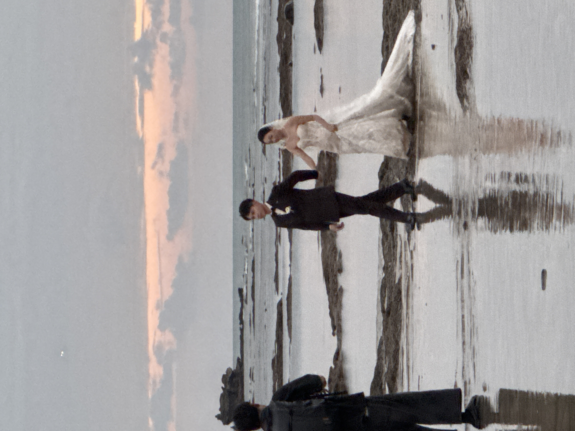 An overcast, late-afternoon shot on a rocky shoreline captures a wedding party posing for photos. A photographer stands on the left, facing away from the camera, his back to the viewer. He is dressed in black and wears a backpack. To his right, a bride and groom walk hand-in-hand across the wet sand. The groom, in a dark suit and bow tie, leads the way. The bride, in a strapless white wedding gown with a long train, follows. In the background, waves crash against a rocky outcrop, and the sky is a muted grey with streaks of soft pink and orange light from the setting sun. The sea is calm, with reflections of the sky and the figures on the shore. The image is a candid shot, suggesting a moment captured during a photoshoot at a beach location in Kuta Selatan, Indonesia.