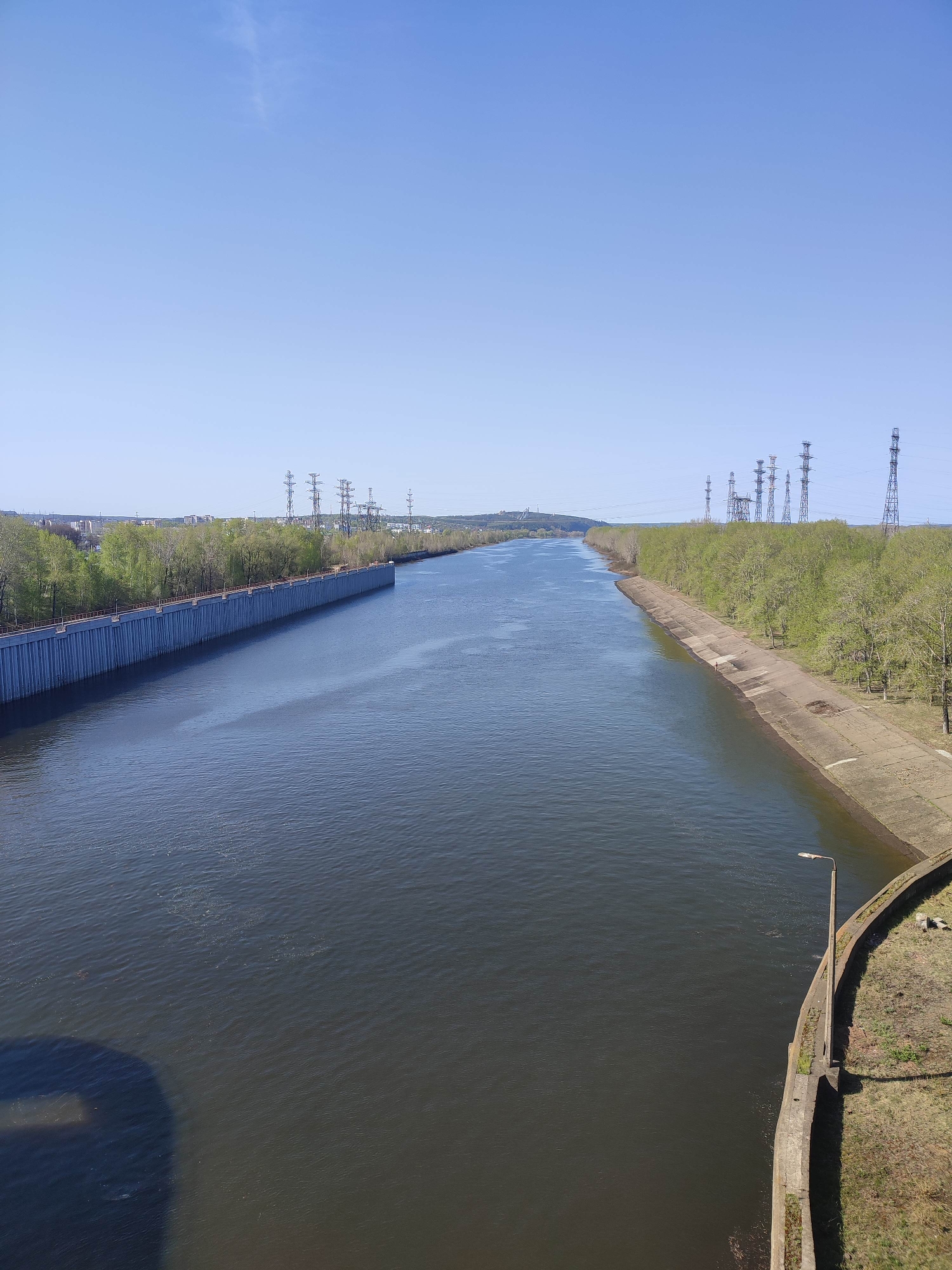 The image depicts a long, wide canal under a clear blue sky. The canal is flanked on both sides by concrete embankments. On the left side, the embankment transitions into a vertical wall. On the right side, a street lamp is visible. Power lines and towers are visible in the distance. The location appears to be a city, possibly named Unknown. The vegetation indicates it may be springtime. There are no people visible.