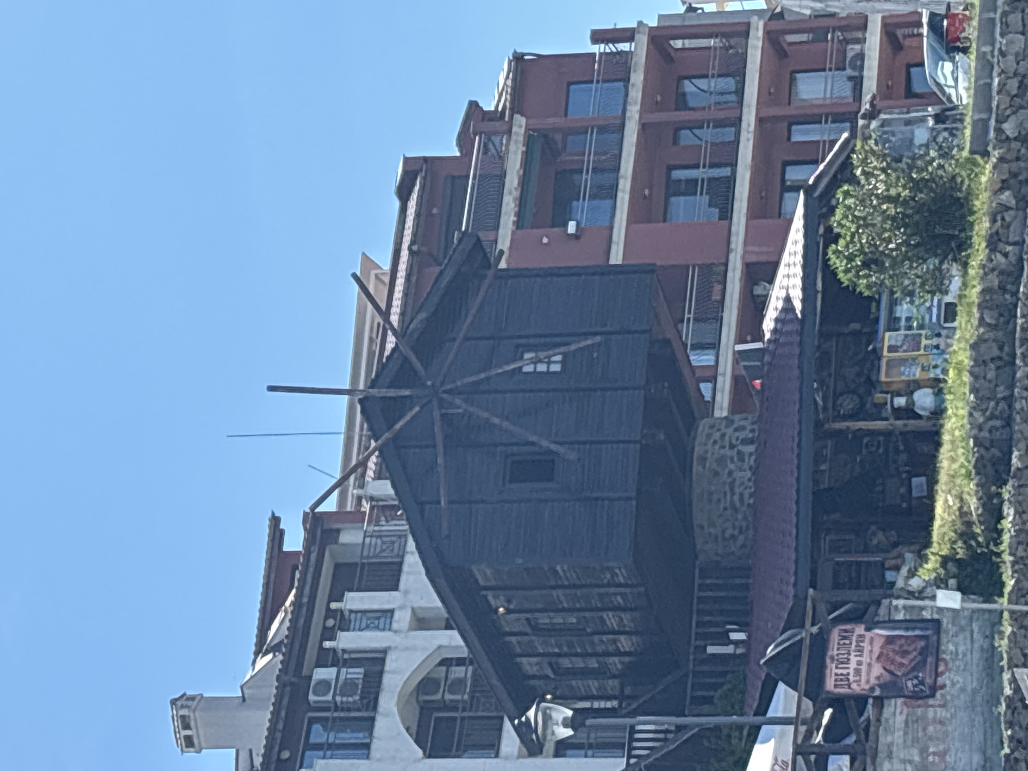 A close-up, eye-level outdoor shot captures a dark wooden, novelty windmill structure perched atop a stone base, set against a backdrop of multi-story buildings. The windmill is angled slightly to the left, its dark wood contrasting with the bright, clear blue sky. The building to the left is white with multiple balconies and air conditioning units, while the building to the right is a deep reddish-brown with a similar balcony structure.
Below the windmill, a red-roofed building with an open-air seating area is visible. A sign hangs from this area, featuring text in Cyrillic alphabet and an image of what appears to be meat. A lamppost stands to the left of the windmill. In the foreground, a low stone wall borders a grassy area with a small bush. The overall scene suggests a commercial area, possibly a tourist destination, on a sunny day. No people are clearly visible.