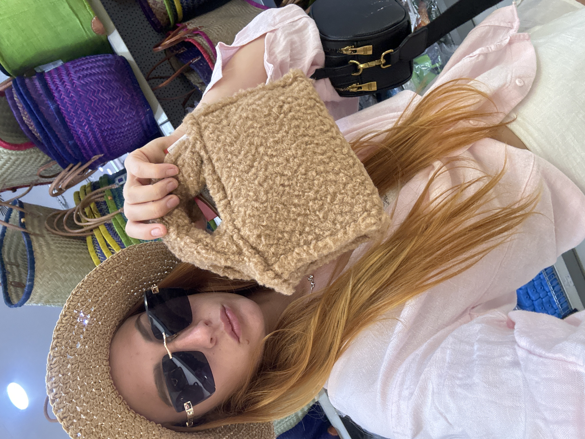 A young woman with long, reddish-blonde hair is taking a selfie. She is wearing a straw hat and black sunglasses. She holds a small, fuzzy, light brown bag in her hands. She is also wearing a light pink, collared shirt. In the background, there are various colorful woven baskets and a black cylindrical bag with gold zippers. The image appears to be taken indoors, possibly in a market or shop, given the display of baskets. The lighting suggests it could be daytime. There are no visible text elements in the image.