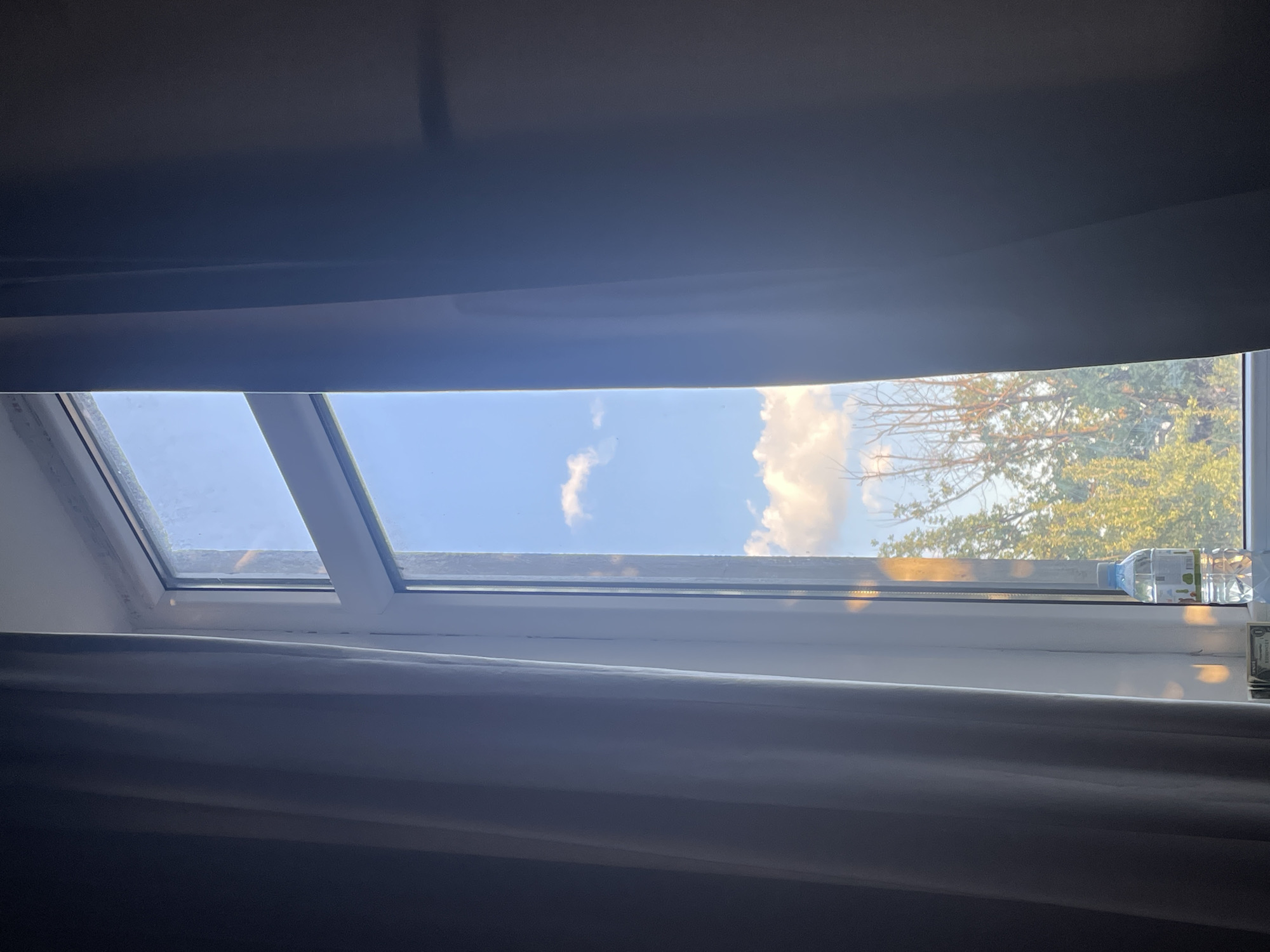 A vertical shot captures a view through a window. The window is framed by dark curtains on both sides, with the right curtain pulled back to reveal the scene. The window itself has a white frame and is divided into sections by a vertical mullion. Outside, the sky is a bright blue, dotted with fluffy white clouds. Sunlight streams in, casting a warm glow on the right side of the window frame and illuminating parts of the trees visible below. The trees appear green and leafy, with some bare branches interspersed. On the windowsill in the lower right corner, there is a clear plastic bottle of water with a white cap and a label. In front of the bottle, there are some folded paper currency bills, possibly money. The overall impression is of a calm, bright day, viewed from indoors. The image does not contain any specific landmarks that would definitively place it in Almaty, Kazakhstan.