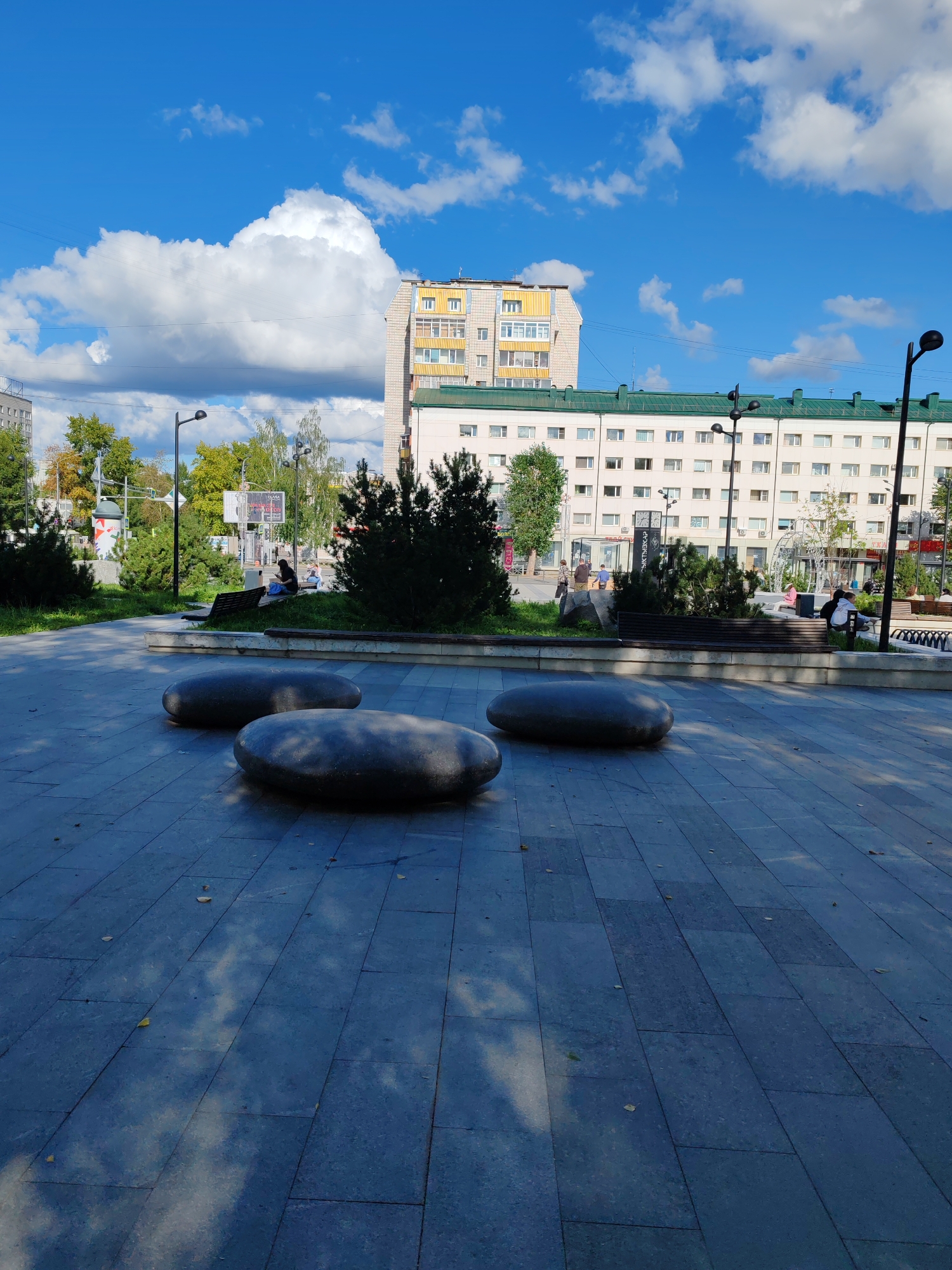 The image shows a public plaza in Syktyvkar, Russia, on a sunny day with scattered clouds. In the foreground, three large, smooth, oval-shaped stone sculptures are arranged on a tiled pavement. The pavement is made of dark gray rectangular tiles, some of which are reflecting the light and shadows of the clouds overhead.
In the middle ground, a park area features green grass, trees, and benches. Several people are seated on benches or walking in this area. Behind the park, a multi-story building with a light-colored facade and a green roof dominates the background. There are windows on the building's facade, and some air conditioning units are visible. A taller building with a yellow section at the top is visible to the left of the main building. Streetlights and some signage are also present in the scene. The sky is bright blue with fluffy white clouds.