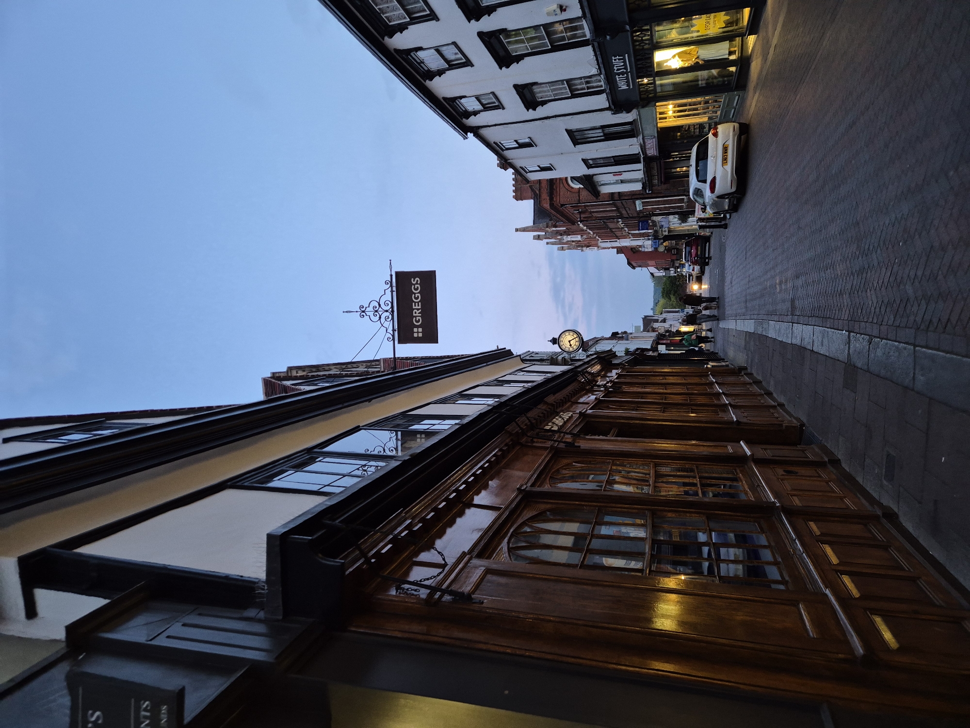 The photograph, taken from a low angle and looking upwards, captures a narrow, brick-paved street in what appears to be a historic town in West Suffolk, United Kingdom. Buildings line both sides, creating a sense of enclosure.
On the left, a multi-story building features a distinct dark wooden facade on its lower levels, with prominent arched windows and intricate detailing. Its upper stories are lighter, possibly timber-framed, with white walls and dark window frames. Hanging prominently from this building is a dark brown sign with the white text "GREGGS". Further along this side, a traditional clock face mounted on a decorative bracket is visible.
On the right side of the street, another building with a white upper facade and dark-framed windows houses a "WHITE STUFF" store, indicated by a prominent sign above its illuminated windows. Further down the street, the architecture continues with a mix of brickwork and older styles.
Artificial lights from shop windows cast warm glows, particularly on the wooden facade, suggesting it is either dusk or evening. The sky above is a clear, light blue, indicating fading daylight or an overcast day. A white car with the license plate "LM10 WMT" is parked on the right side of the street. A few blurry figures of pedestrians can be seen walking further in the distance. Partial, largely unreadable text, possibly including "S. NTS" and "NNN", is visible on a sign at the bottom left edge of the frame. The overall atmosphere is quiet and evocative of a traditional British market town at twilight.