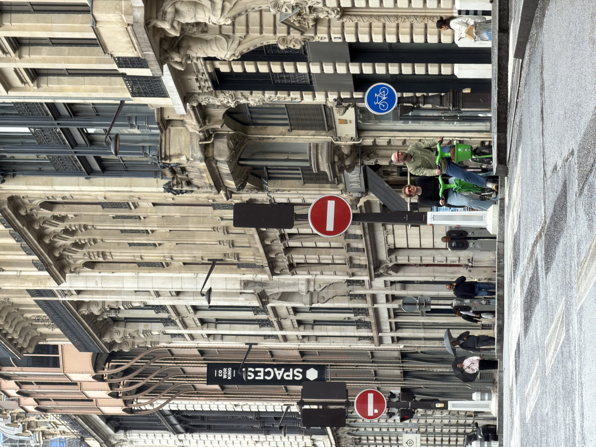 The image depicts a street scene in Paris, France, with an emphasis on the architectural details of the buildings lining the street. The ornate facades are characterized by classical designs and decorative elements.
Several pedestrians are visible on the sidewalk:
- Two individuals ride green scooters.
- Others walk along the sidewalk, with one using an umbrella.
Street signs and traffic signals are interspersed throughout the scene, including bicycle route sign, "no entry" signs, and a sign for "Spaces - Co Working". The street surface is wet.