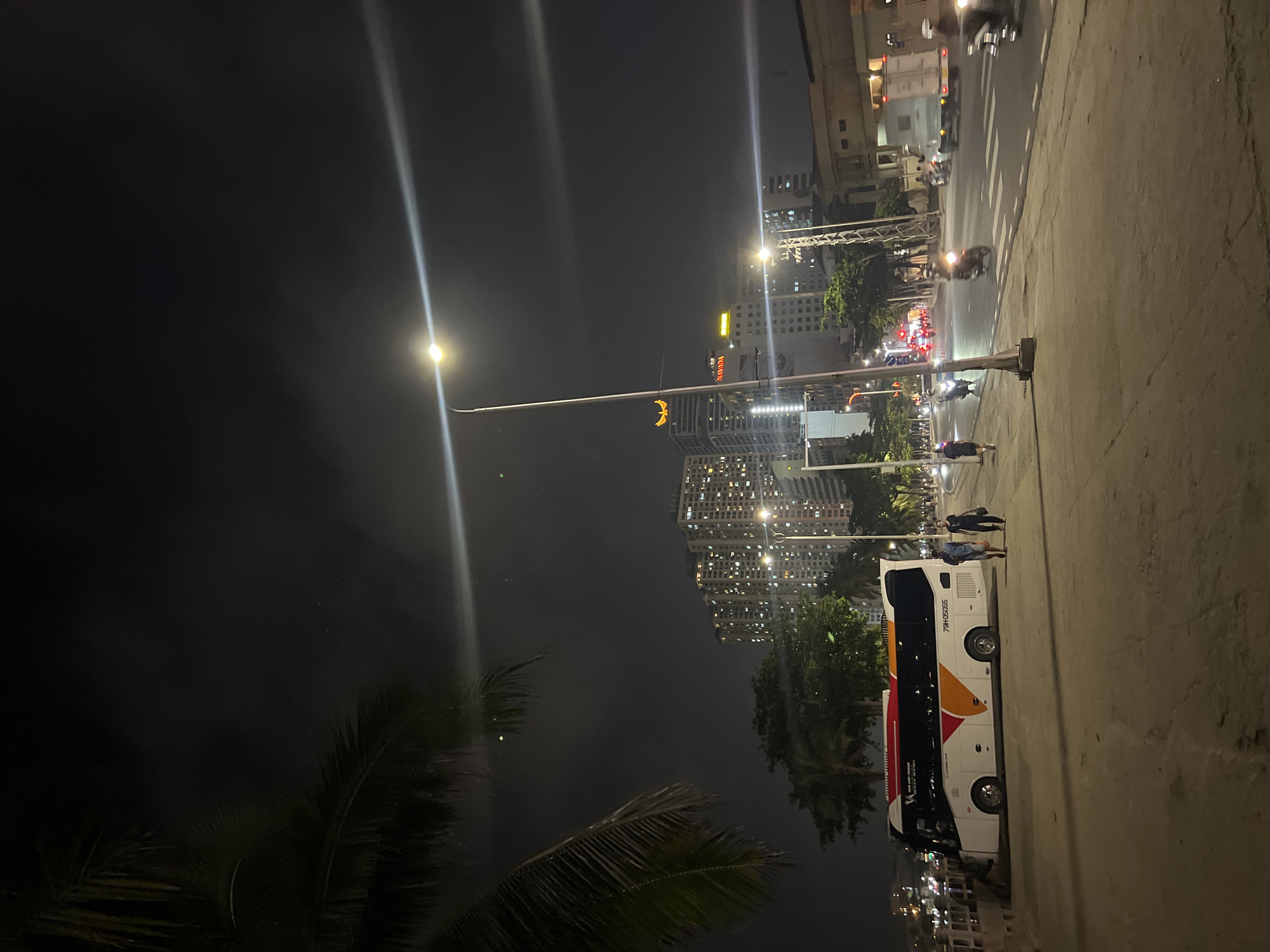 The photograph shows a nighttime scene in Bắc Nha Trang, Vietnam. Tall buildings with lighted windows dominate the skyline. A street with traffic is visible, lit by streetlights. A large white bus with red and orange graphics is parked along a sidewalk. Several people are standing and walking near the bus. The foreground includes palm trees against the dark sky and water. Streetlights cast long streaks of light upward.