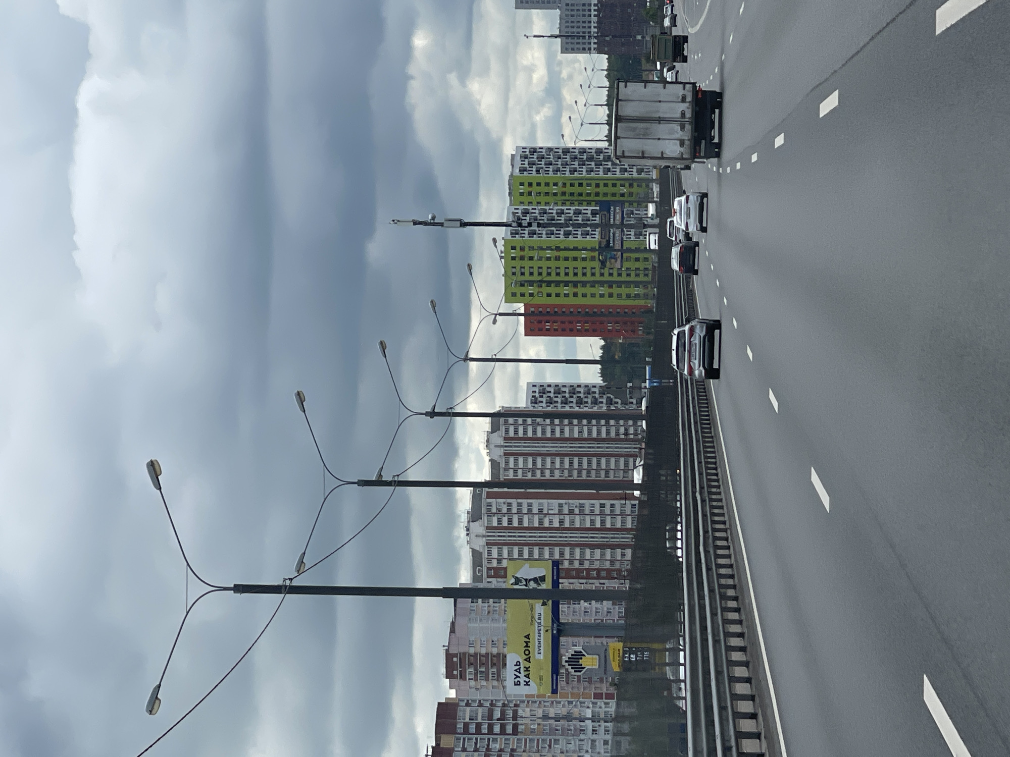The image shows a multi-lane highway, likely an overpass, with traffic moving along it. The setting is in Novodrozhzhino, Russia, as indicated by the Russian text on a large advertising banner visible alongside the highway. The sky is overcast, with gray clouds dominating the upper portion of the picture.
Several tall apartment buildings are visible in the background, painted in different colors (red, green, white, etc.) and arranged in rows. Streetlights line the side of the highway. Cars, a truck, and a van are traveling on the highway.