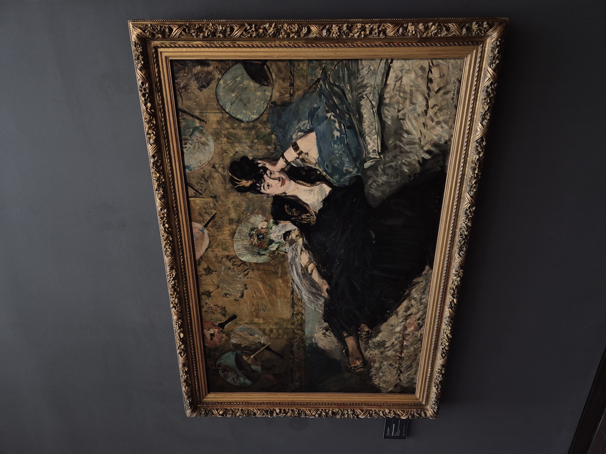 The image displays a painting by Édouard Manet titled "Olympia" within a golden, ornate frame. The painting depicts a nude woman, Olympia, reclining on a couch with a black servant presenting her with flowers. The background features wallpaper with a floral pattern and an oriental screen. The painting is displayed on a dark gray wall, likely in a museum. The lighting is subdued, highlighting the artwork. The perspective is angled, showing the painting from a slight side view, with the bottom left corner of the frame closest to the viewer. A small information plaque is visible to the left of the painting. The artwork is characteristic of Impressionist art, with visible brushstrokes and a focus on everyday subjects. The scene is static and posed, typical of formal portraiture, yet Manet's depiction of Olympia was considered controversial for its frankness and realism.