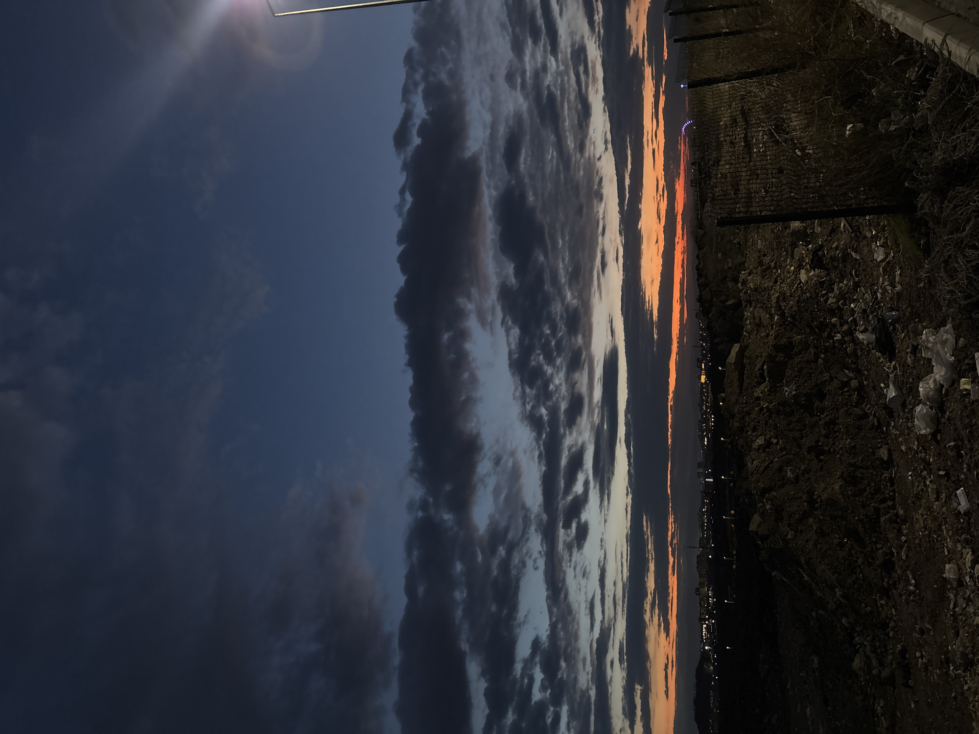 The image captures a twilight scene, possibly a sunset, in Yerevan, Armenia. The sky dominates the view with dark, textured clouds on the left transitioning to orange and white hues near the horizon on the right. A light source, possibly a street lamp, is visible at the top left, causing lens flare. Below the horizon, faint lights suggest the presence of the city skyline. The foreground contains a dark, uneven terrain with a wire fence running along its edge, indicating a border between the urban and natural landscape. No people are identifiable in the image.