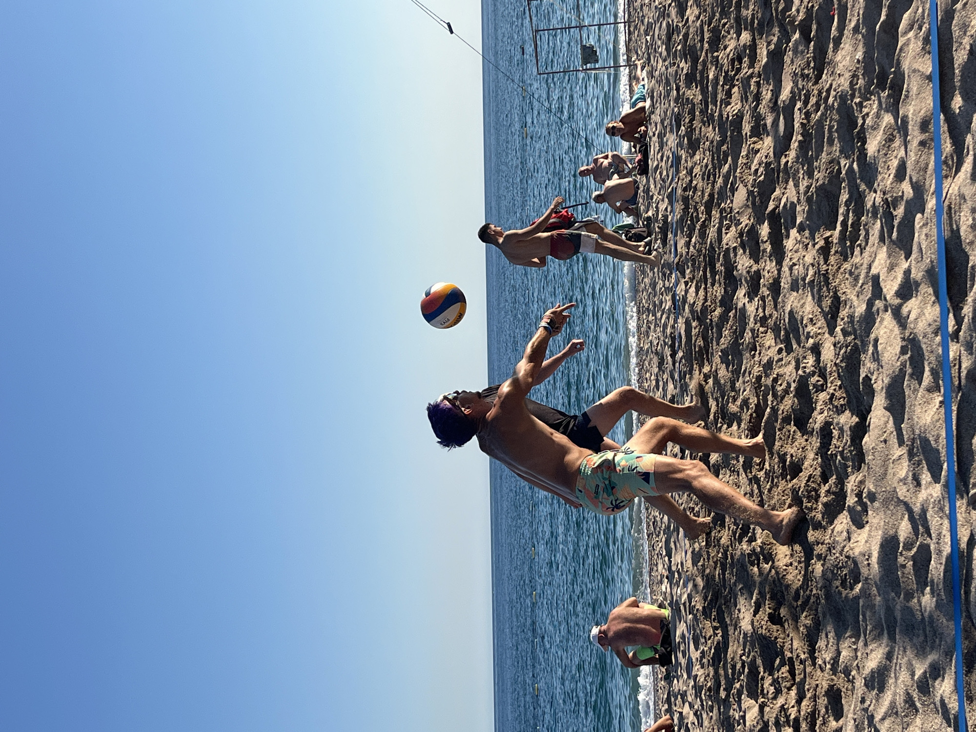 A group of men are playing beach volleyball on a sandy beach in Zankovići, Montenegro. The sky is clear and blue, and the ocean is visible in the background. The men are wearing swim trunks and are in various stages of playing the game. One man is mid-air, hitting the volleyball, while another is preparing to receive it. Other people are sitting and lying on the beach, watching the game. The sand is the primary ground surface, with a blue line marking the volleyball court boundary.