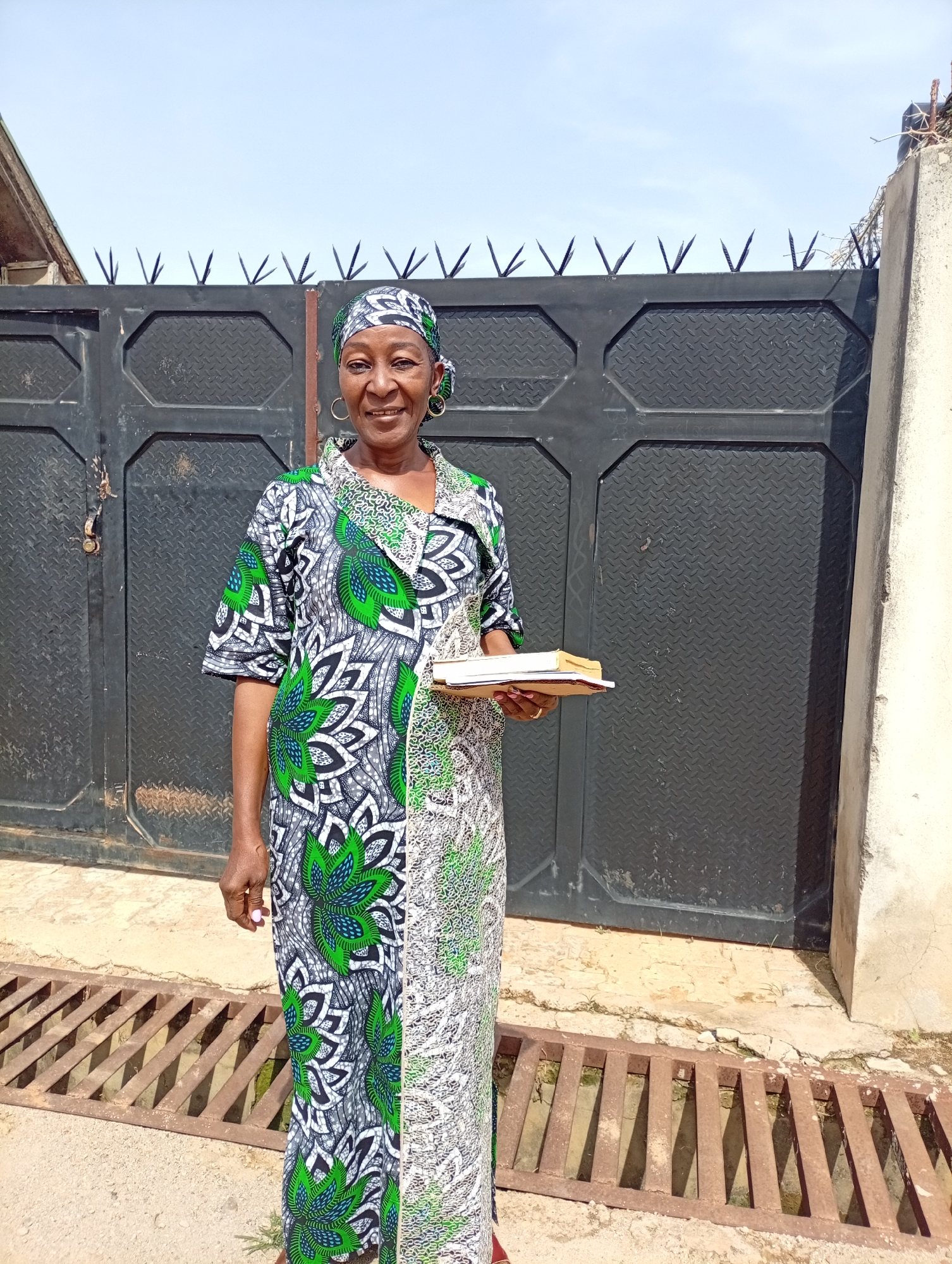 An eye-level, medium shot presents a middle-aged Black woman standing in front of a dark gray metal gate topped with spikes, which is set in a light gray concrete wall. The location is identified as Unknown, Unknown.
The woman wears a long, patterned dress in shades of green, white, and gray. Her head is wrapped in a matching headscarf. She also has circular earrings. She holds a stack of books in her left hand. Her facial expression is one of a smile, with her eyes looking toward the camera.
Beneath the gate, in the foreground, is a rusty, metal drainage grate set into a light-colored concrete surface. The background is a clear, bright sky. Part of the roof of a building is visible to the left of the gate.