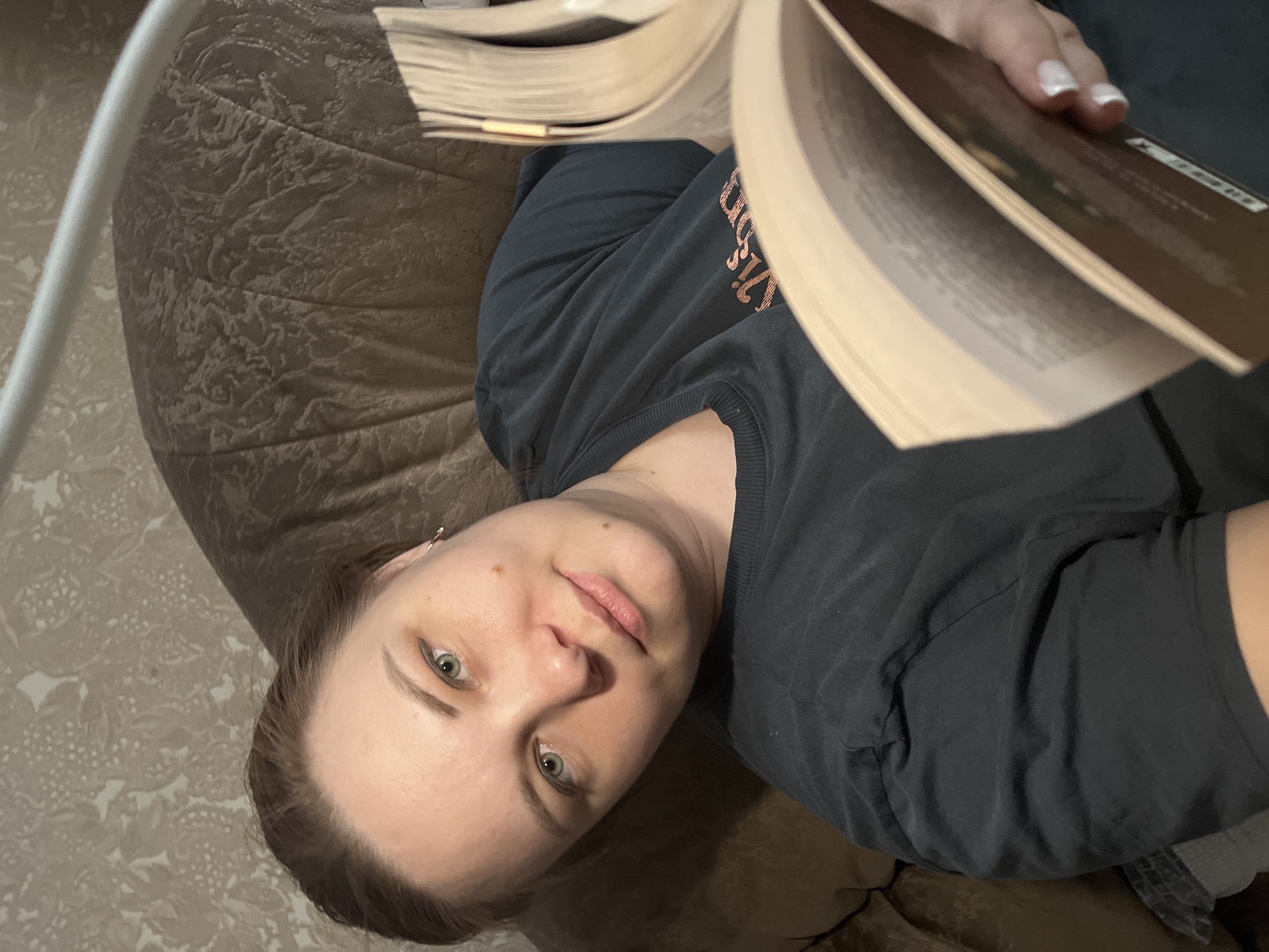A woman is reclining on a brown, patterned cushion or beanbag while reading a book. She is wearing a dark gray t-shirt. The environment appears to be indoors, against a wall with floral wallpaper. The city of Usman, Russia, is not verifiable from the context of this image.