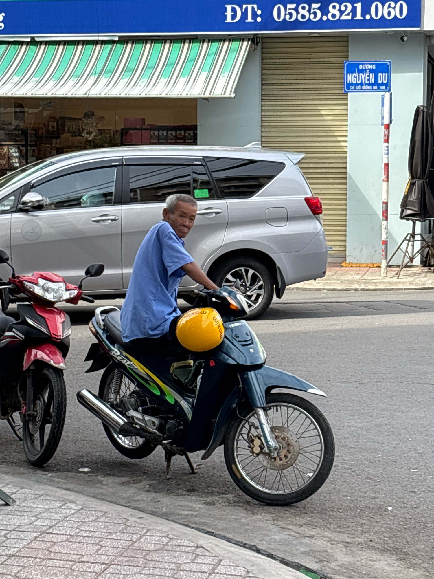 An elderly Vietnamese man is sitting on a dark blue motorcycle, wearing a blue short-sleeved shirt. He is looking towards the camera with a slight smile. A bright yellow helmet with black text is on the seat of his motorcycle. To his left, a red and black motorcycle is parked. In the background, a silver minivan is visible, parked on the street. The street is paved, with a tiled sidewalk on the left side of the image. A building with a blue awning displaying the phone number "ĐT: 0585.821.060" is behind the vehicles. A street sign in Vietnamese that reads "ĐƯỜNG NGUYỄN DU" is also visible on the right side of the image, indicating the location is Nguyen Du Street in Nha Trang, Vietnam. The man is on a street in Nha Trang, Vietnam.