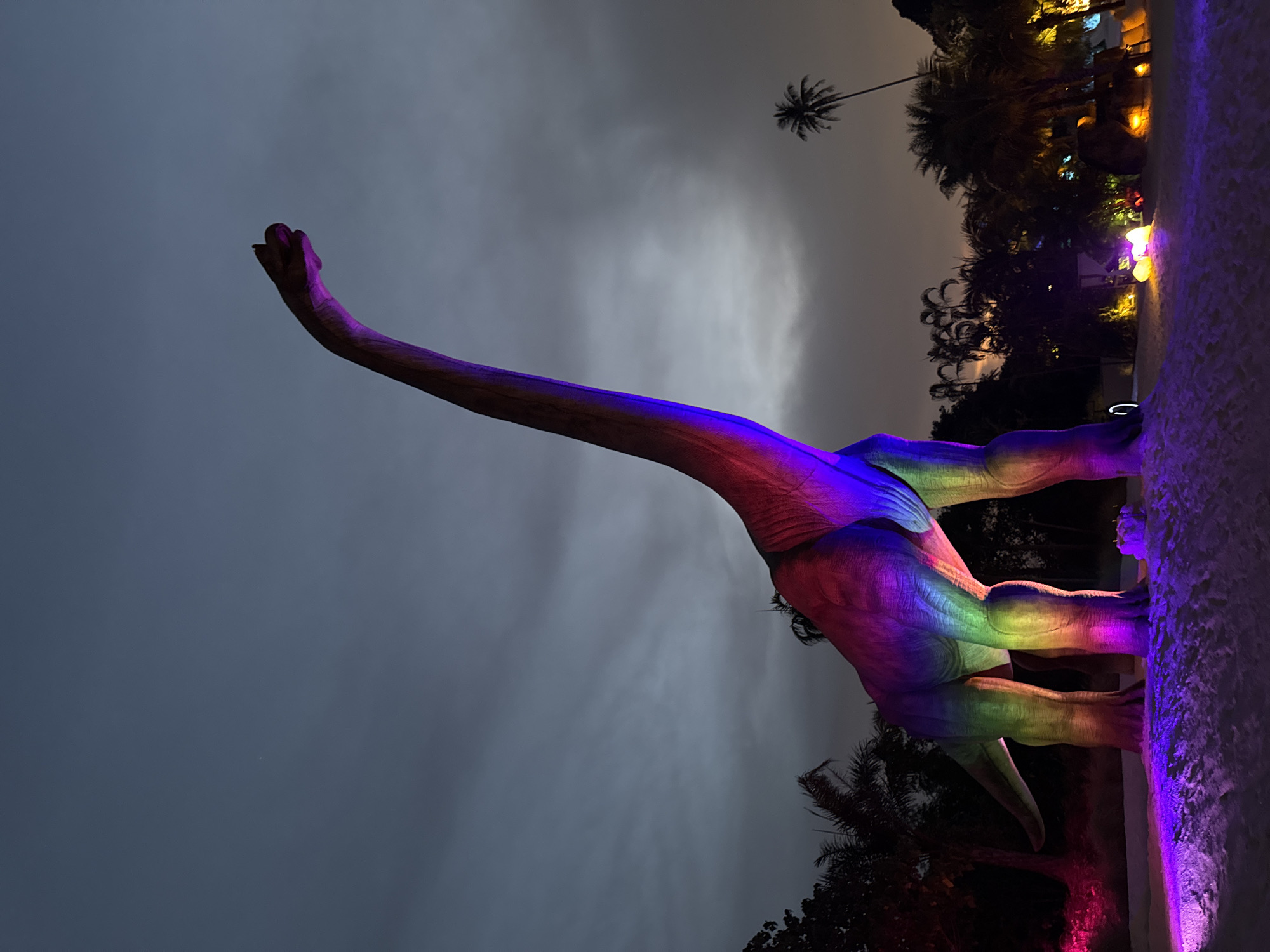 This image depicts a striking outdoor scene at night or late dusk in Baan Lipa Noi, Thailand, likely within a park or attraction. The central subject is a colossal dinosaur statue, resembling a Brachiosaurus or Apatosaurus, brightly illuminated with a gradient of artificial rainbow colors. Its long neck stretches gracefully upwards towards the overcast, dark grey sky, with its head pointing towards the upper left of the frame.
The statue's coloration transitions from purple and blue on its raised neck and head, through vivid reds, oranges, yellows, and greens along its body, and down to purples and pinks on its robust legs. The material of the statue appears textured and solid.
In the background, to the right, a dense cluster of dark tropical trees and foliage is visible, with various light sources casting soft yellow, orange, and purple glows, hinting at further illuminated features. A tall, slender palm tree stands silhouetted against the cloudy sky near the upper right, reinforcing the tropical environment. Along the bottom edge of the frame, a light-colored, textured pathway or low wall is bathed in purple light. The sky is heavily clouded, indicating an evening setting. The scene is static, showcasing an impressive and colorful display. No visible text is present.