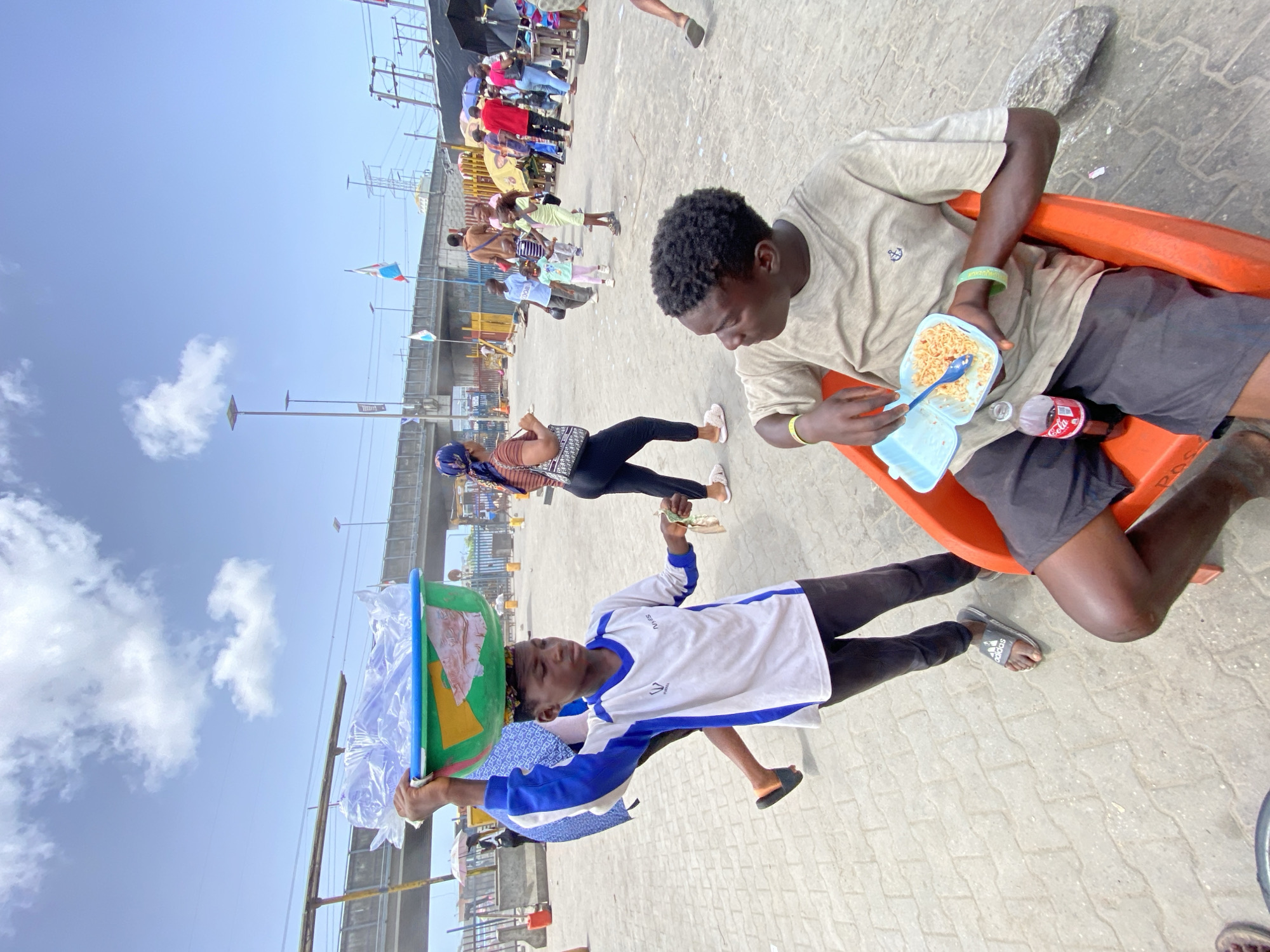 The image depicts an outdoor scene on a paved surface in what is inferred to be Lekki, Nigeria based on context clues. A young man sits on an orange plastic chair, eating noodles from a blue plastic container with a blue spoon. A bottle of Coca-Cola is next to him. Another young man balances a blue and green container filled with plastic-wrapped items on his head. A woman in black leggings and a patterned shirt is walking in the background. In the distance, a crowd of people is visible near a bridge and several flags, including the Nigerian flag. The sky is blue with scattered clouds.