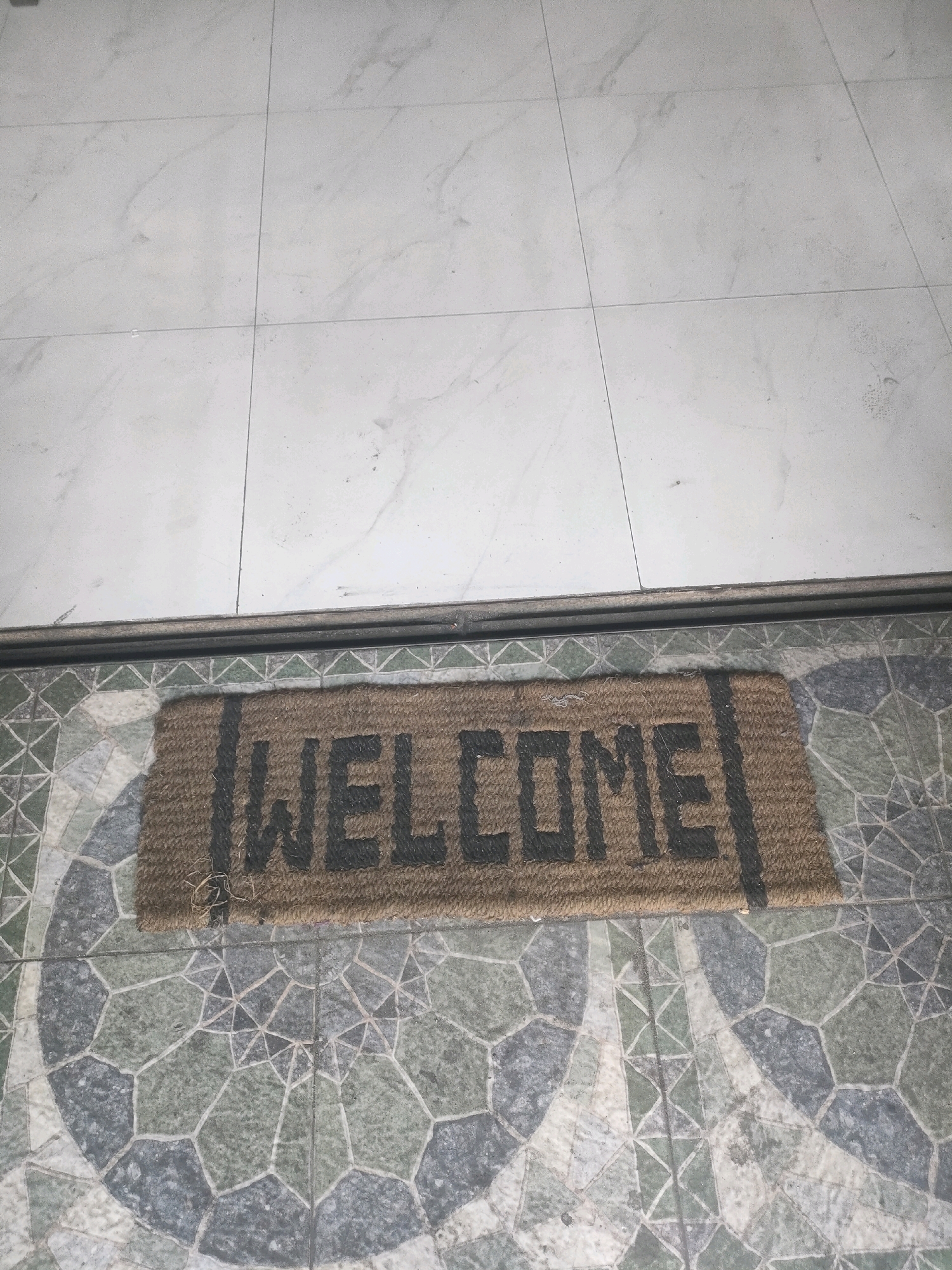 A coir doormat with the word "WELCOME" in black letters is placed on a floor with patterned tiles. Above the doormat, the floor transitions to larger, lighter-colored tiles. The scene is indoors, likely at the entrance of a residential or commercial building in Surabaya, Indonesia. There are no people or visible actions in the image.