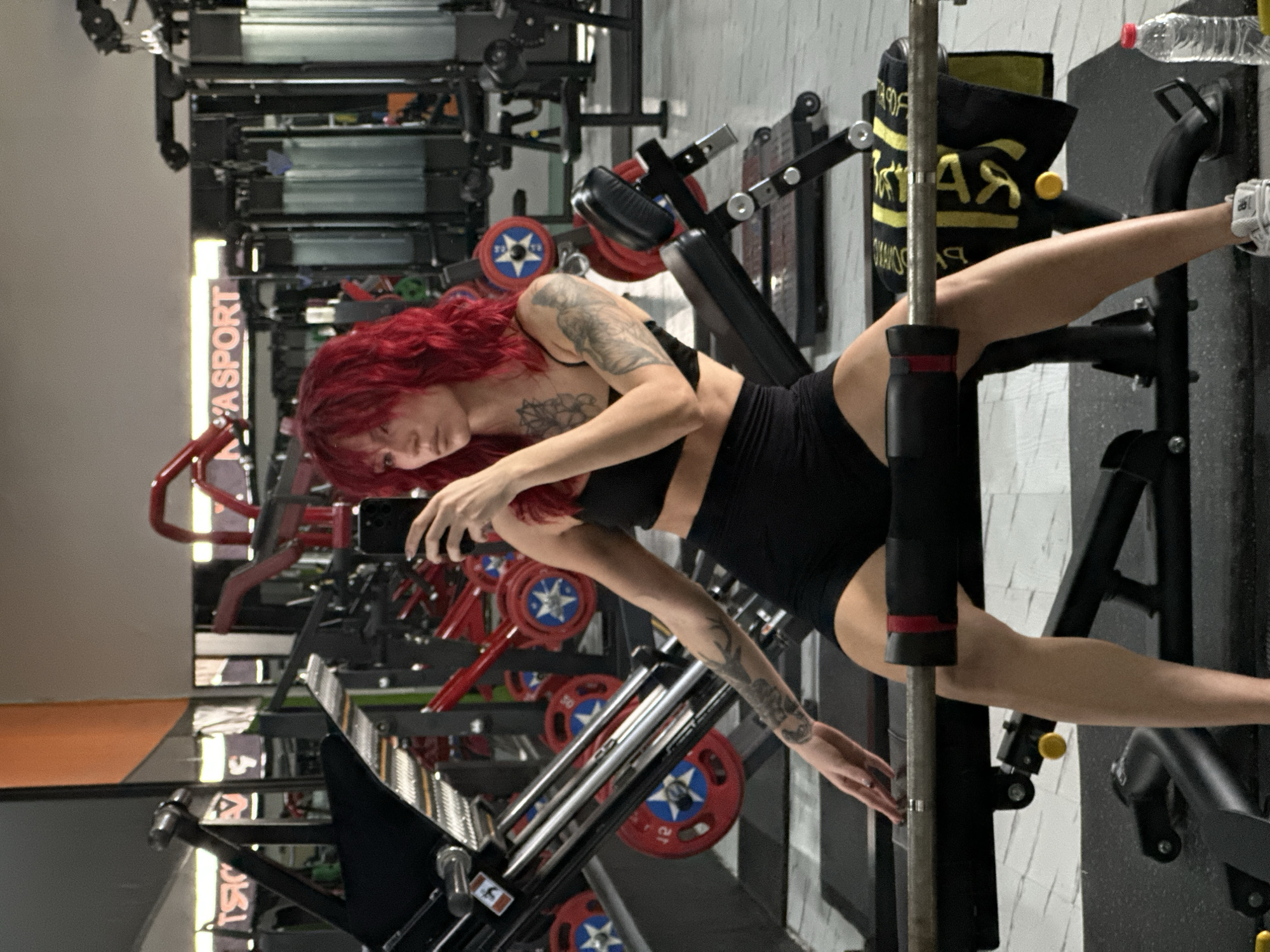 A woman with vibrant red hair is taking a selfie in a gym. She is wearing a black sports bra and black shorts, and is seated on a weight bench with a barbell across her legs. Her arms are tattooed, and she is holding a black smartphone in her right hand. The gym is equipped with various weight machines and equipment, reflected in the mirrors behind her. There is a water bottle on the floor to the right of the image. The overall environment appears to be modern and functional. The location, North Kuta, Indonesia, is not visually discernible from the image content itself, but is provided as contextual information.