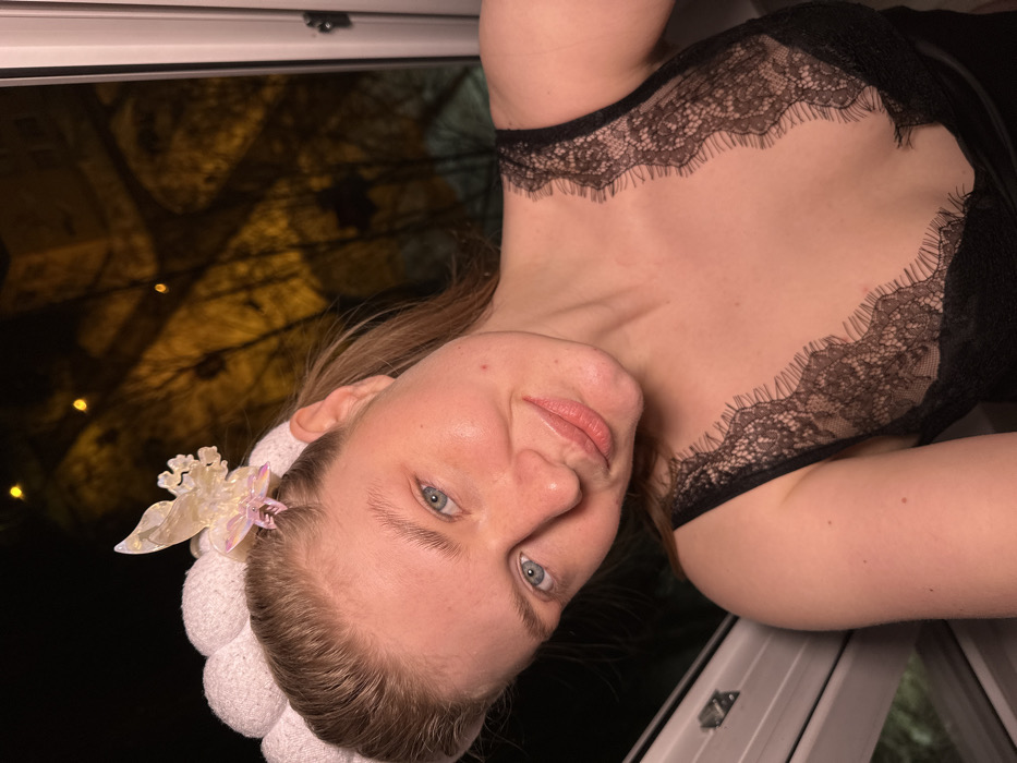 An indoor portrait captures a fair-skinned adult female, appearing in her 20s or 30s, at night in Kazan, Russia. She has light brown hair and blue eyes, looking directly at the camera with a slight smile. She wears a white, textured headband. A translucent, iridescent hair clip, shaped like a butterfly or similar intricate design, is attached to the headband above her right temple. Her attire includes a black, V-neck top with extensive lace detailing on the straps and décolletage. The subject is positioned in front of a window, identifiable by a white frame visible along the left and bottom edges. The exterior background is dark, indicating night, with blurred, warm orange-yellow lights in the distance. Silhouettes of bare tree branches are discernible against the ambient light. A faint, dark architectural silhouette, consistent with an urban building, is visible in the upper right background.
