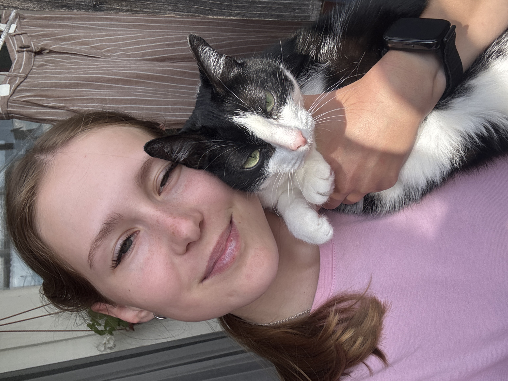 The image captures a young adult female holding a black and white tuxedo cat in what appears to be an indoor or enclosed balcony setting in Chaikovsky, Russia.
The female, with light complexion and light brown hair, wears a plain light pink crew-neck t-shirt and a silver chain necklace. A small silver hoop earring is visible on her left ear, and a dark-faced smartwatch with a black fabric strap is secured on her right wrist. She exhibits a subtle smile towards the viewer, her eyes slightly narrowed, suggesting a response to bright light.
She cradles the cat with her right arm, holding it close to her chest. The cat, identifiable by its black fur with white markings on its chest, belly, and paws, has distinct green eyes, a pink nose, and long white whiskers. Its eyes are partially squinted, and its ears are slightly flattened, contributing to a somewhat neutral or contemplative expression.
The background on the left features a white vertical wall with a hanging green potted plant that displays small white flowers. On the right, partially visible, are light brown trousers with thin white vertical stripes, suspended on a clothes hanger against a darker backdrop, indicative of a domestic interior. Strong, natural light illuminates the scene from the left, creating distinct shadows across the woman's arm and the cat's fur.