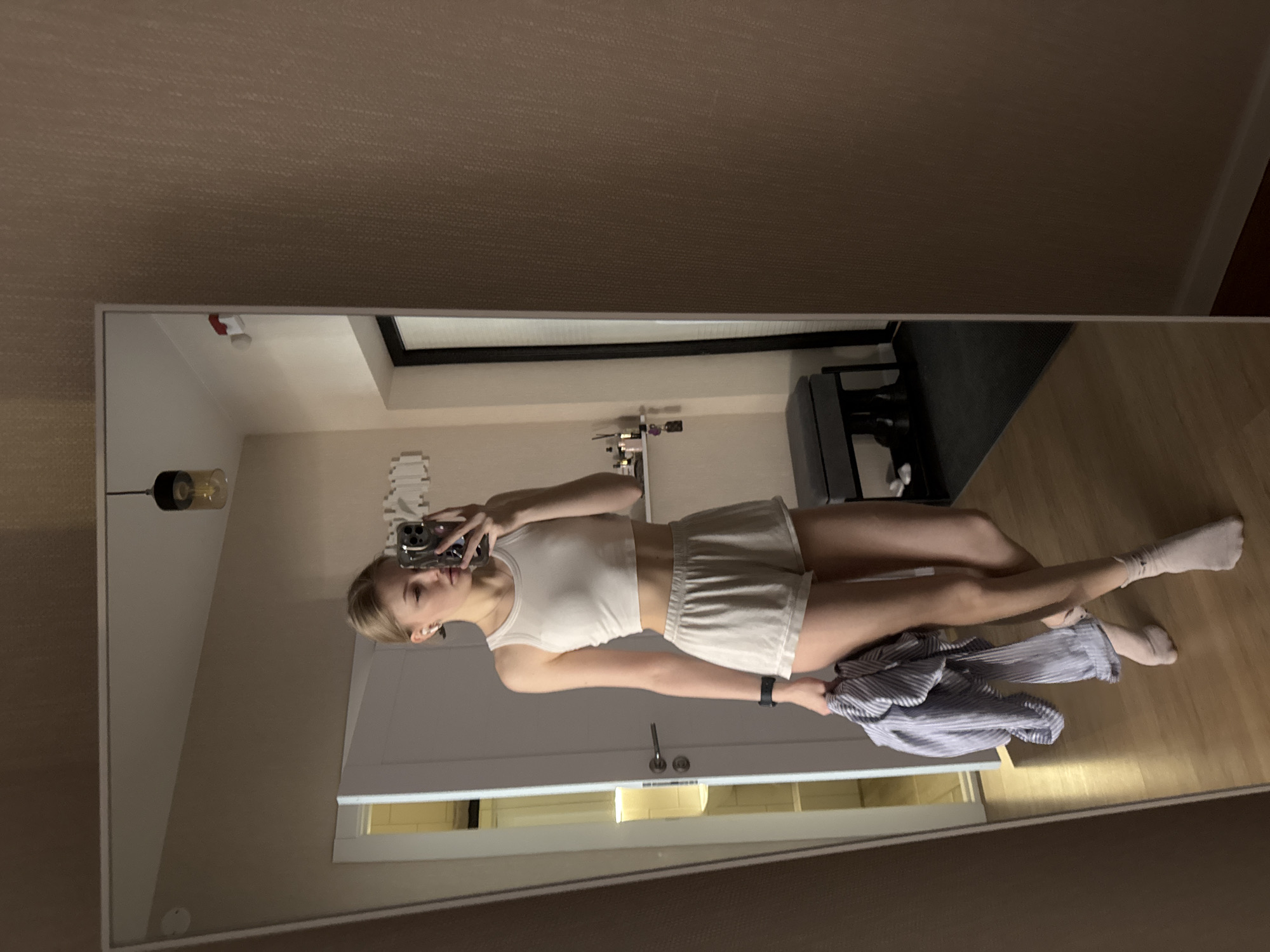 A young woman in a white top and shorts is reflected in a full-length mirror. She holds a phone in her right hand and a striped garment in her left. She wears white socks. The room behind her features a closed white door, light wood flooring, a fitness machine, and storage. Location is not discernible.