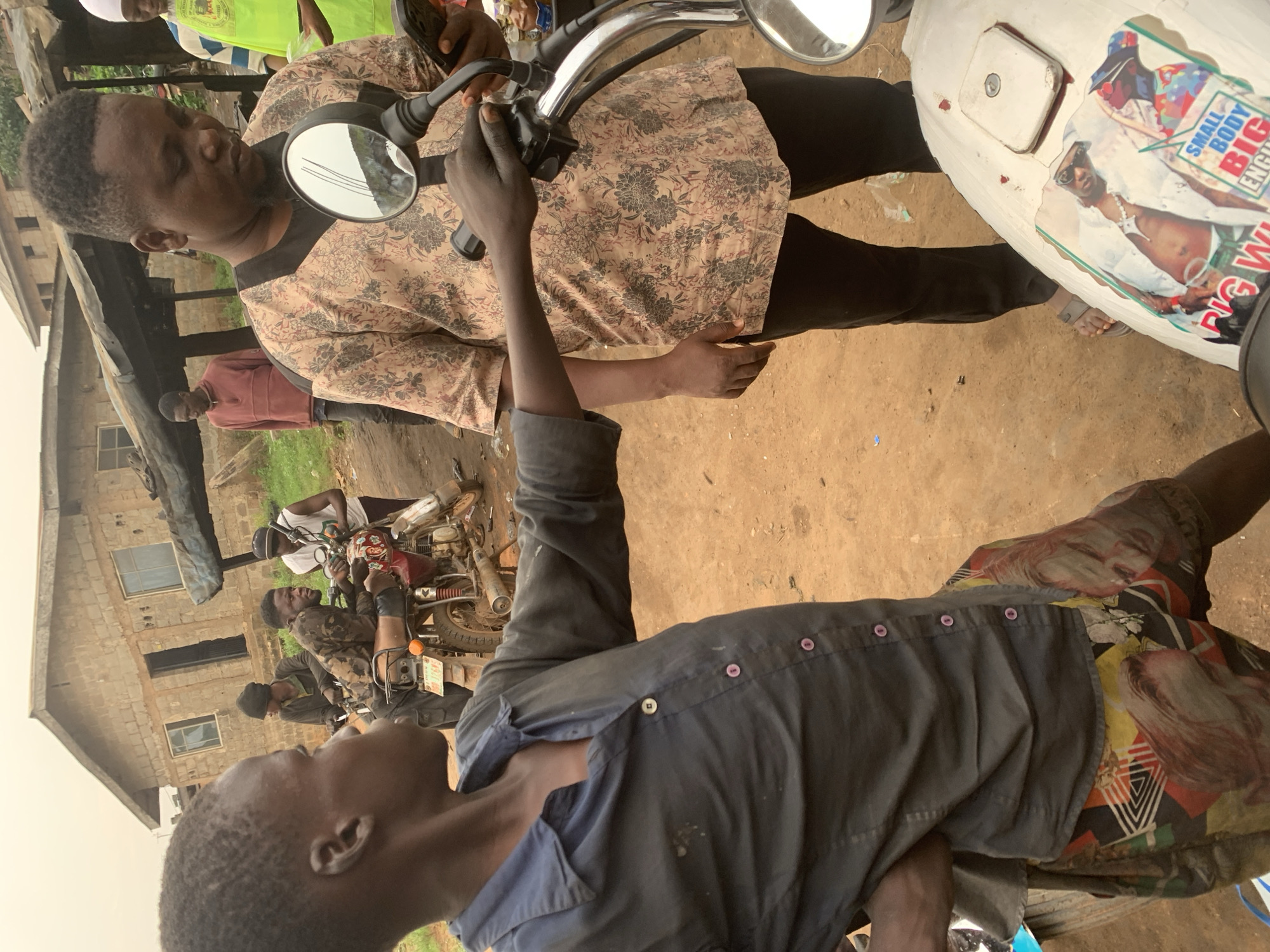 In Shagamu, Nigeria, a young boy in a dark blue collared shirt and patterned shorts stands to the left of the frame, facing right and interacting with a man in a patterned shirt and black pants. The man, holding a phone in his right hand, is looking down at it. A motorcycle is in the foreground, with a sticker on its fuel tank featuring a person wearing sunglasses and a vest. In the background, other individuals are visible near a building with a corrugated metal roof, and a second motorcycle is parked. The ground is dirt, and the overall environment appears to be an outdoor marketplace or roadside area.