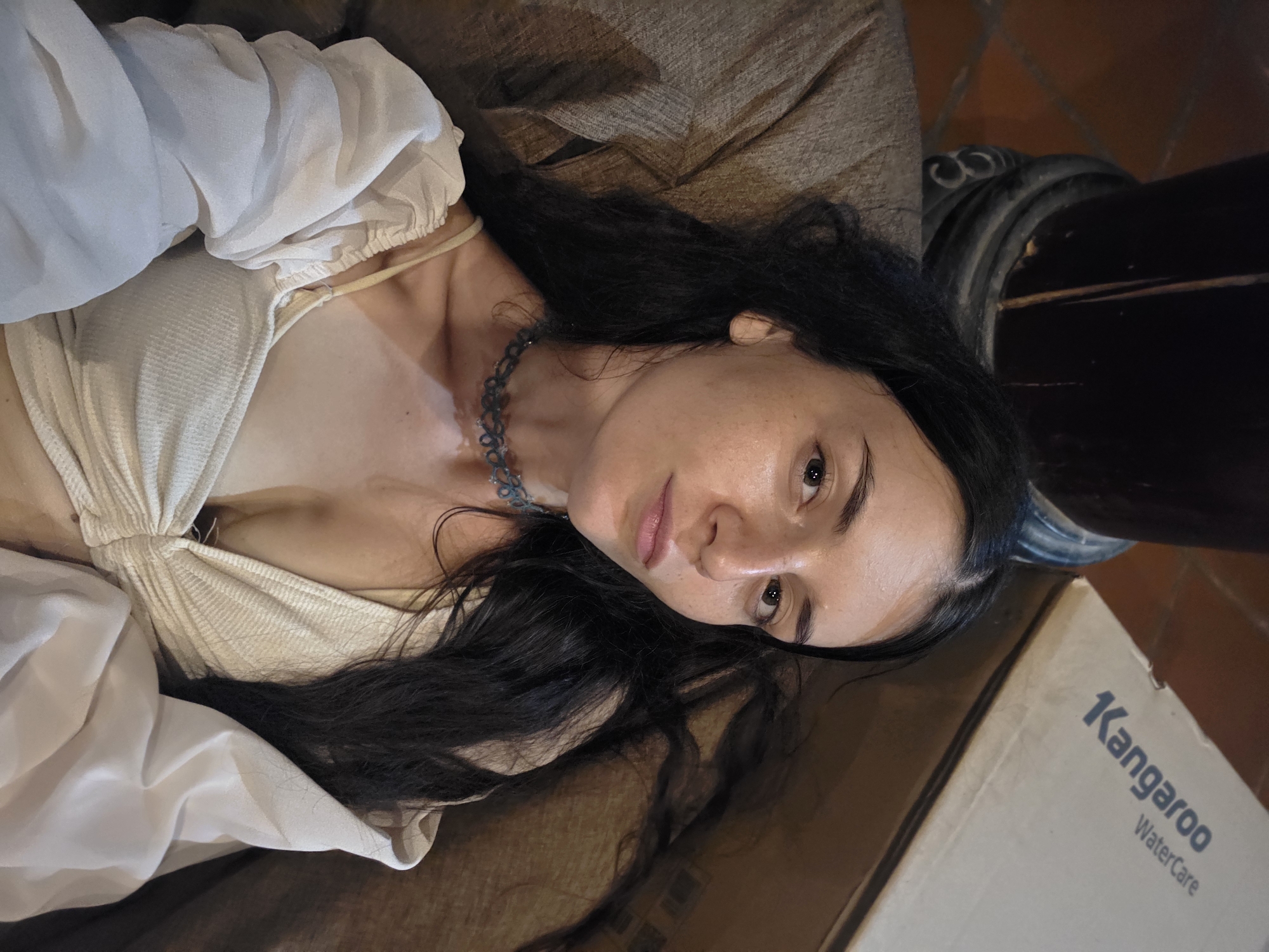 The image captures a woman lying down, looking directly at the camera with a neutral, direct gaze. She has long, dark, wavy hair that spreads around her head. Her eyes are dark brown, and her complexion is fair, with some lighter patches visible on her chest and neck. She is wearing a light, possibly off-white or beige, sleeveless top with a gathered or ruched design on the bodice. A voluminous, puffy sleeve in a similar light shade is visible on her left side. Around her neck, she wears a delicate, blue-grey choker-style necklace.
She is positioned on a brown, textured surface, likely a sofa or cushion. To the lower right, a light brown cardboard box is visible with the brand "Kangaroo" prominently displayed in blue capital letters, with "WaterCare" written below it. In the background, behind her head and the box, there is a dark, possibly wooden or metal, structure with curved details, and a reddish-brown tiled surface. The scene is indoors, suggesting a relaxed setting, potentially in Mũi Né, Vietnam. The lighting is soft, creating an intimate atmosphere.