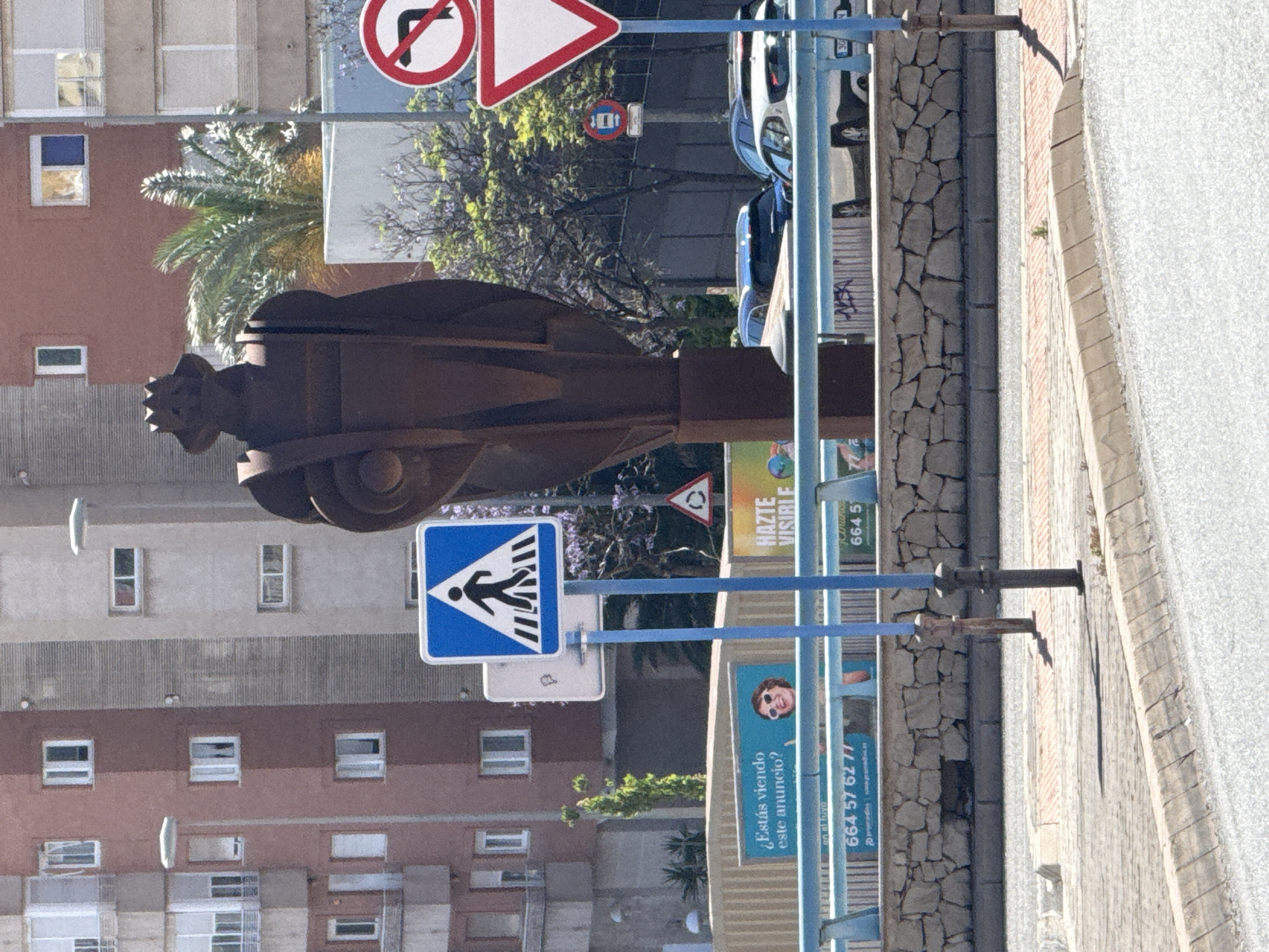 The image depicts an outdoor urban scene in Unknown, Unknown. A weathered metal sculpture of an abstract, potentially human figure stands prominently in the foreground. Behind the sculpture is a multi-story residential building with a red and gray facade and evenly spaced windows. Traffic signs, including a pedestrian crossing sign and a "no right turn" sign, are visible, attached to blue metal posts. Adjacent to the sculpture is a panel advertising services with a phone number. A street with light gray stone pavement runs along the right side of the image. The background includes green trees and a partially visible car. The overall image is captured in daylight and suggests a typical street scene in a city.