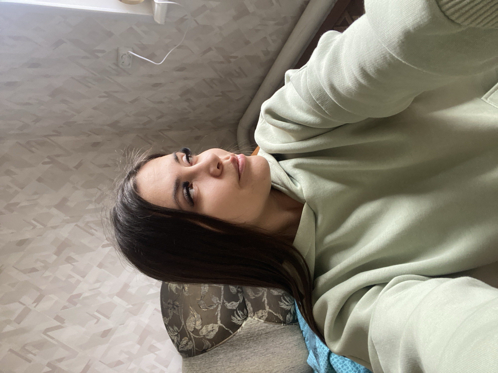 The image shows a young woman with long, dark hair, depicted from a low-angle perspective. Her head is tilted slightly back as she looks upwards and towards the right side of the frame with a neutral, possibly contemplative expression. She is wearing a light pastel green hooded sweatshirt, with the hood down. Her dark hair cascades over her shoulder and against what appears to be the armrest of a piece of furniture.
This furniture, likely a sofa or armchair, is upholstered in a dark fabric with a subtle, reflective floral or leaf pattern. A piece of blue patterned fabric, possibly a blanket or cushion, is visible near her left arm and side.
The background consists primarily of a light-colored wall with a textured wallpaper pattern, characterized by interlocking geometric shapes or abstract lines in varying shades of light grey and off-white. In the upper portion of the image, an electrical outlet with a white cable plugged into it is visible on the wall. Further up, near what appears to be the ceiling line, a light fixture or vent can be seen. The overall setting suggests an indoor, domestic environment, likely a living room or bedroom. There is no visible text. The general lighting suggests it is daytime. The location context is Zhigalki, Russia.