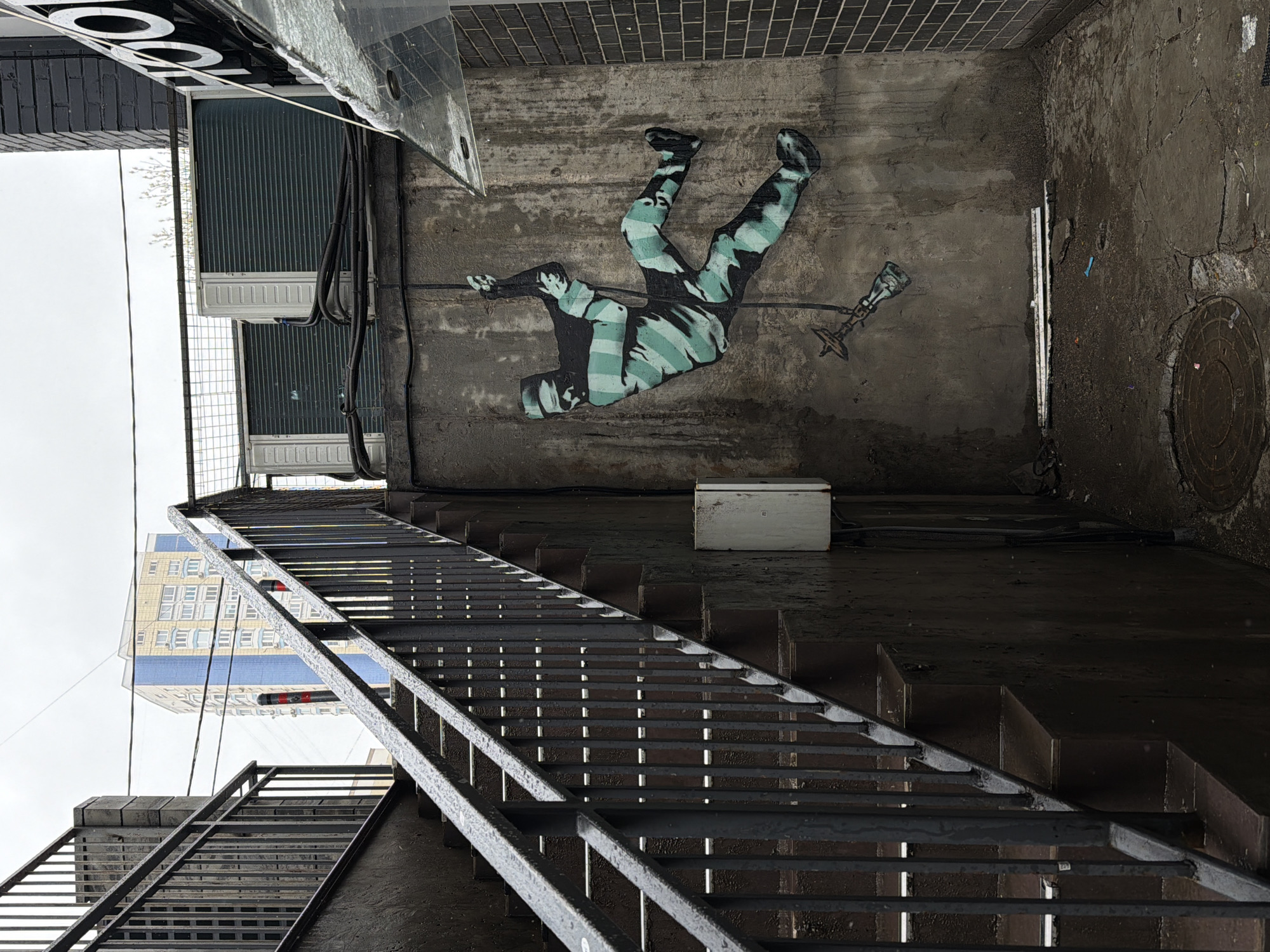 The image, when correctly oriented, displays a damp and grimy urban alleyway or service area under an overcast sky, characteristic of an environment like Moscow, Russia. A prominent mural on the upper right-hand concrete wall features a stylized figure, rendered in a teal and dark grey camouflage pattern, appearing to fall or be suspended. The figure holds an object resembling a torch or a spear with decorative elements.
A dark, wet metal staircase, likely a fire escape, extends diagonally from the lower left foreground towards the mid-right, its gridded steps and railings showing moisture. The ground beneath the staircase and stretching across the lower right is dark, wet concrete, where a circular metal manhole cover is visible. A small white electrical box is mounted on the wall near the base of the mural.
On the left side of the scene, two large, rectangular air conditioning condenser units are mounted on an adjacent building's wall, with multiple black cables running past them and along the ground. Above these units, at the top edge of the image, part of a large, inverted sign showing two "O"s is discernible. In the distance, against the grey sky, a multi-story building with a light-colored facade and numerous windows, along with a distinct blue horizontal stripe, can be seen. The overall impression is one of a utilitarian urban space affected by recent rain or damp conditions.