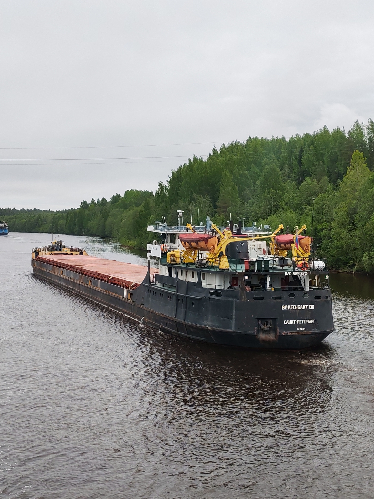 The image depicts a large barge or river cargo vessel, "VOLGO-BALT 136 САНКТ-ПЕТЕРБУРГ", navigating a waterway. The barge is primarily dark in color, with a large, flat deck area covered by an orangish-brown material. There is a smaller vessel further ahead on the water. The surrounding environment consists of lush green trees and vegetation lining both sides of the waterway. The sky is overcast, indicating a cloudy day. The water is dark and reflects the surrounding environment.
The vessel is located on a canal located in the city of Depo, Russia.