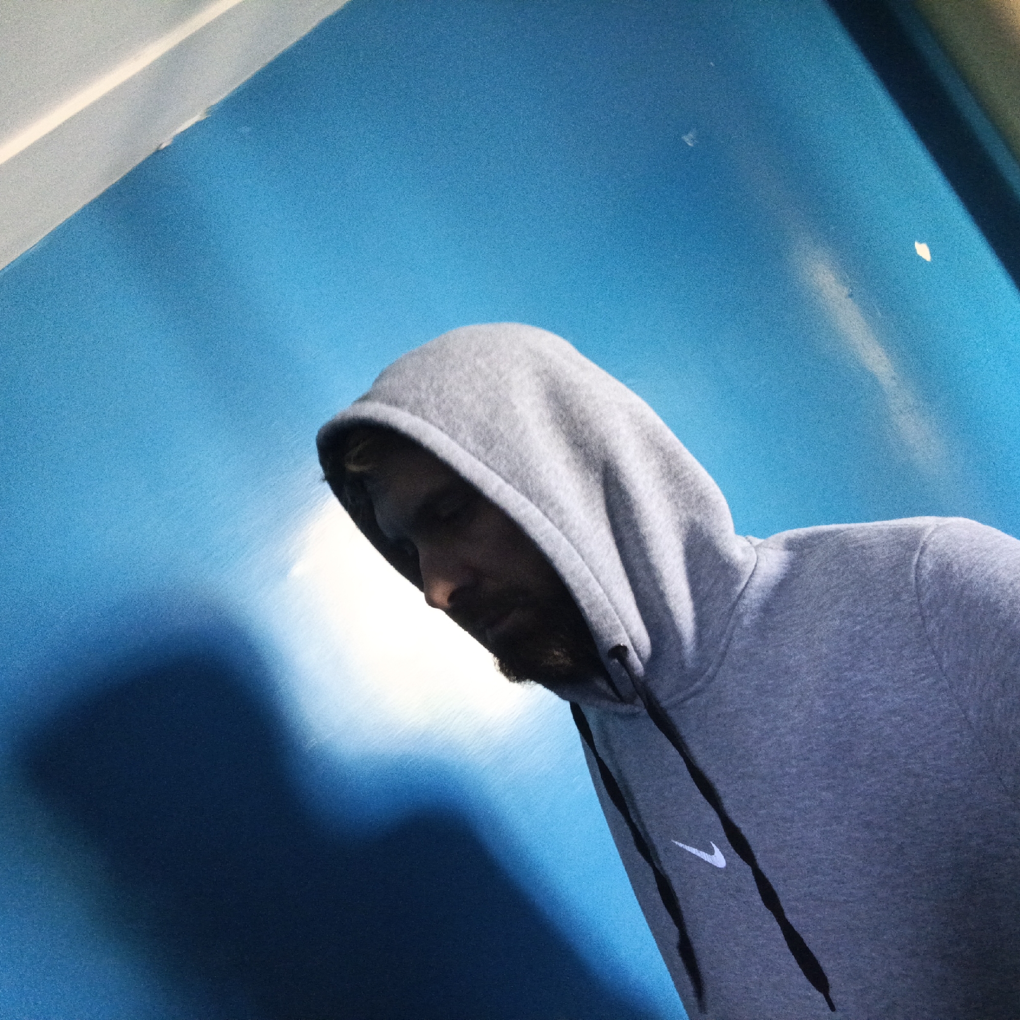 A man in a grey hooded sweatshirt is looking down in front of a blue wall. The man has a beard and the hood is pulled up. The sweatshirt has a white Nike swoosh logo. The background is blurred. It is not possible to tell if this image was taken in Mapkovsky, Russia.