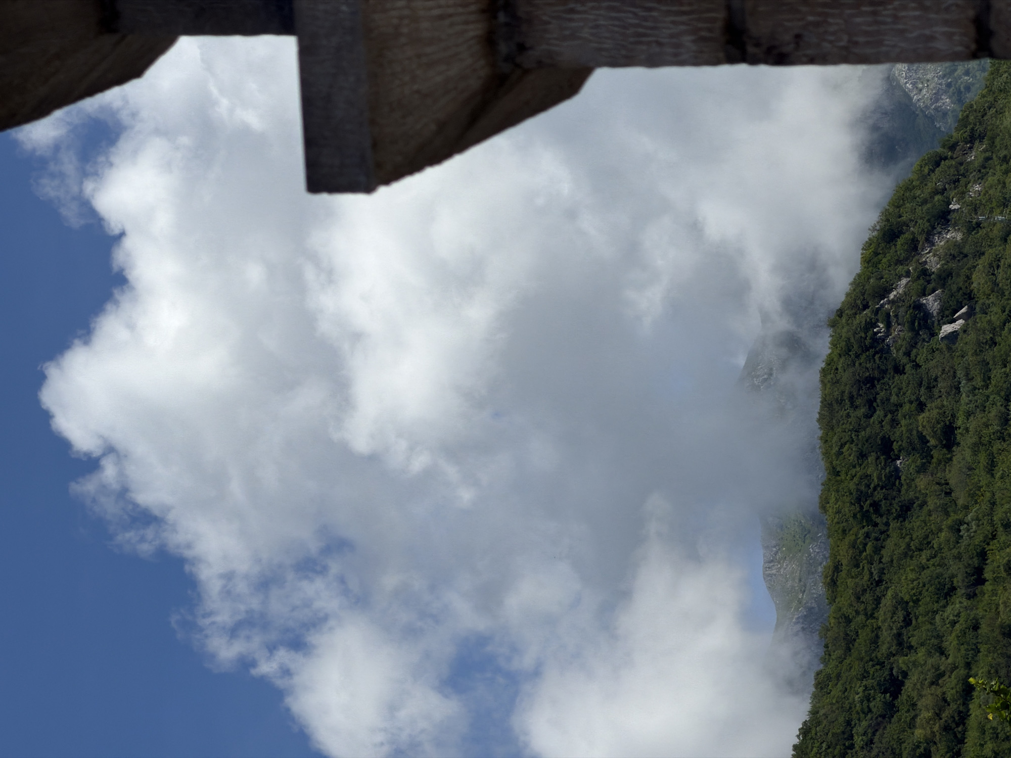The image captures a breathtaking view of a mountainous landscape in Kubasi, Montenegro, seen from a high vantage point. The top of the frame is partially obscured by a dark, textured wooden structure, possibly a beam or a roof edge, suggesting the view is from a building or balcony.
The scene is dominated by a dramatic interplay of natural elements. On the right, a steep, densely forested mountainside descends vertically, covered in lush green trees and rocky outcrops. To its left, a deep valley is engulfed in swirling white and grey mist, which partially conceals the lower parts of distant, rugged mountain peaks. Through the ethereal haze, glimpses of stark, grey rocky cliffs and slopes are visible, hinting at the scale and complexity of the terrain beyond.
The lower-left portion of the image reveals a clear blue sky, interspersed with fluffy, bright white cumulus clouds, indicating fair weather at higher elevations. The contrast between the clear sky and the misty valley suggests dynamic weather conditions typical of mountainous regions.
The time of day appears to be bright daytime, with ample natural light illuminating the clouds and the visible parts of the mountains. There are no visible people, animals, or man-made objects other than the wooden structure in the immediate foreground. No text is visible in the image. The overall impression is one of majestic natural beauty and the vastness of the mountain environment.