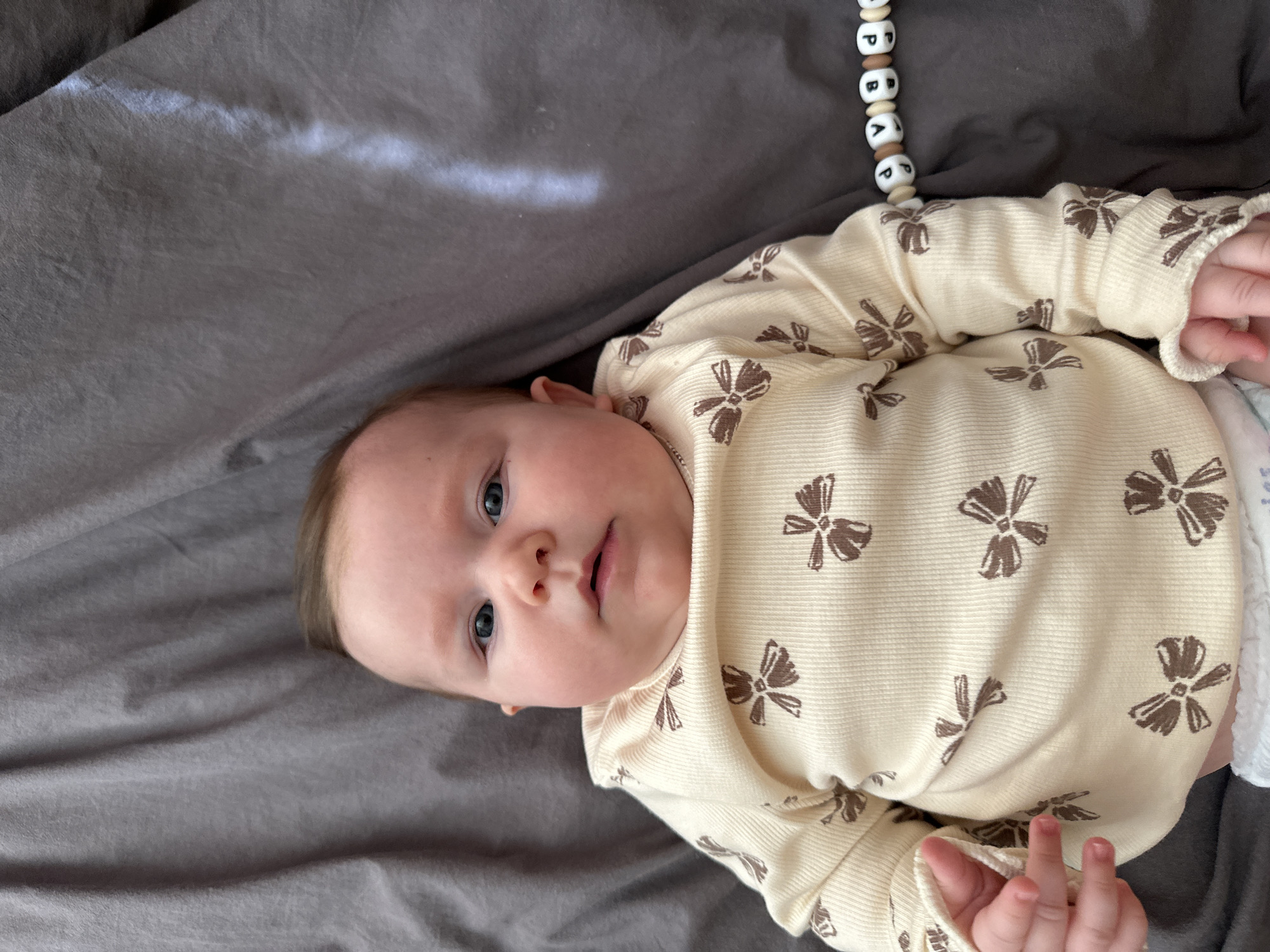 A baby lies on a gray blanket. The baby is wearing a light yellow onesie with brown bow patterns. A string of white and brown beads with letters is visible near the baby's head. The location is not identifiable as Chaikovsky, Russia.