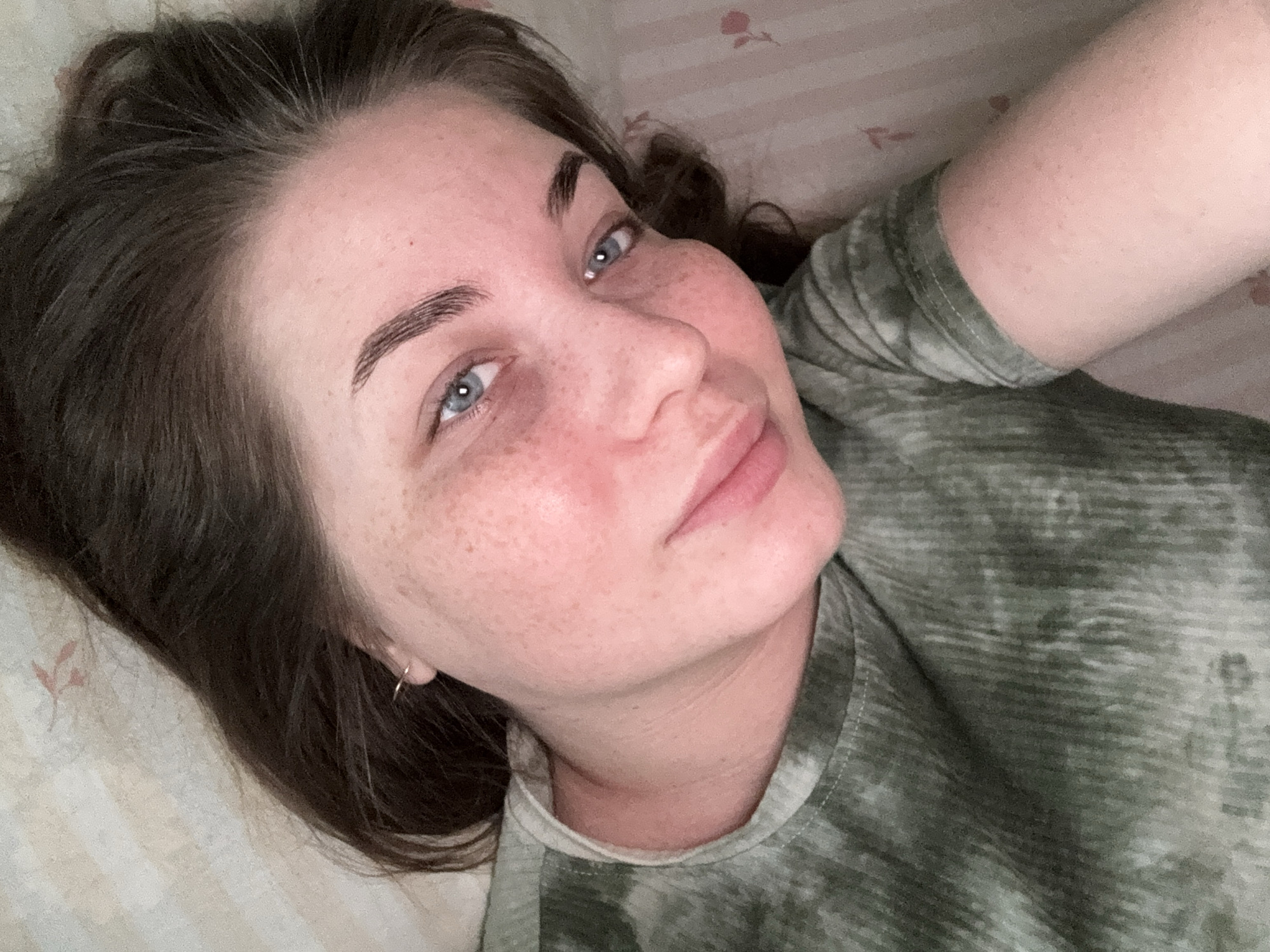 A woman with fair skin, freckles, and blue eyes is lying down and looking at the camera. She has dark brown hair and wears a green t-shirt. She's wearing a small hoop earring in her left ear. The background shows a light-colored bedsheet with a faint floral pattern. The location in the city of Chaikovsky, Russia is unknown, the photograph does not contain this information.