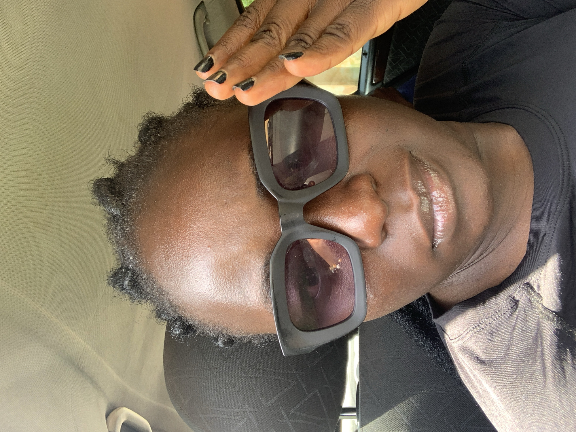 A close-up selfie shows a dark-skinned woman wearing sunglasses and a black t-shirt, sitting in a car. Her right hand is raised, fingers touching her forehead as if shielding her eyes from the sun. The sunglasses are large and black with thick frames. Her hair is styled in small braids. The interior of the car is visible, with the beige headliner and a dark patterned seat in the background. The lighting suggests it is daytime and sunny. The image does not provide any specific details about the city of Jos, Nigeria, nor any context to place the scene there.