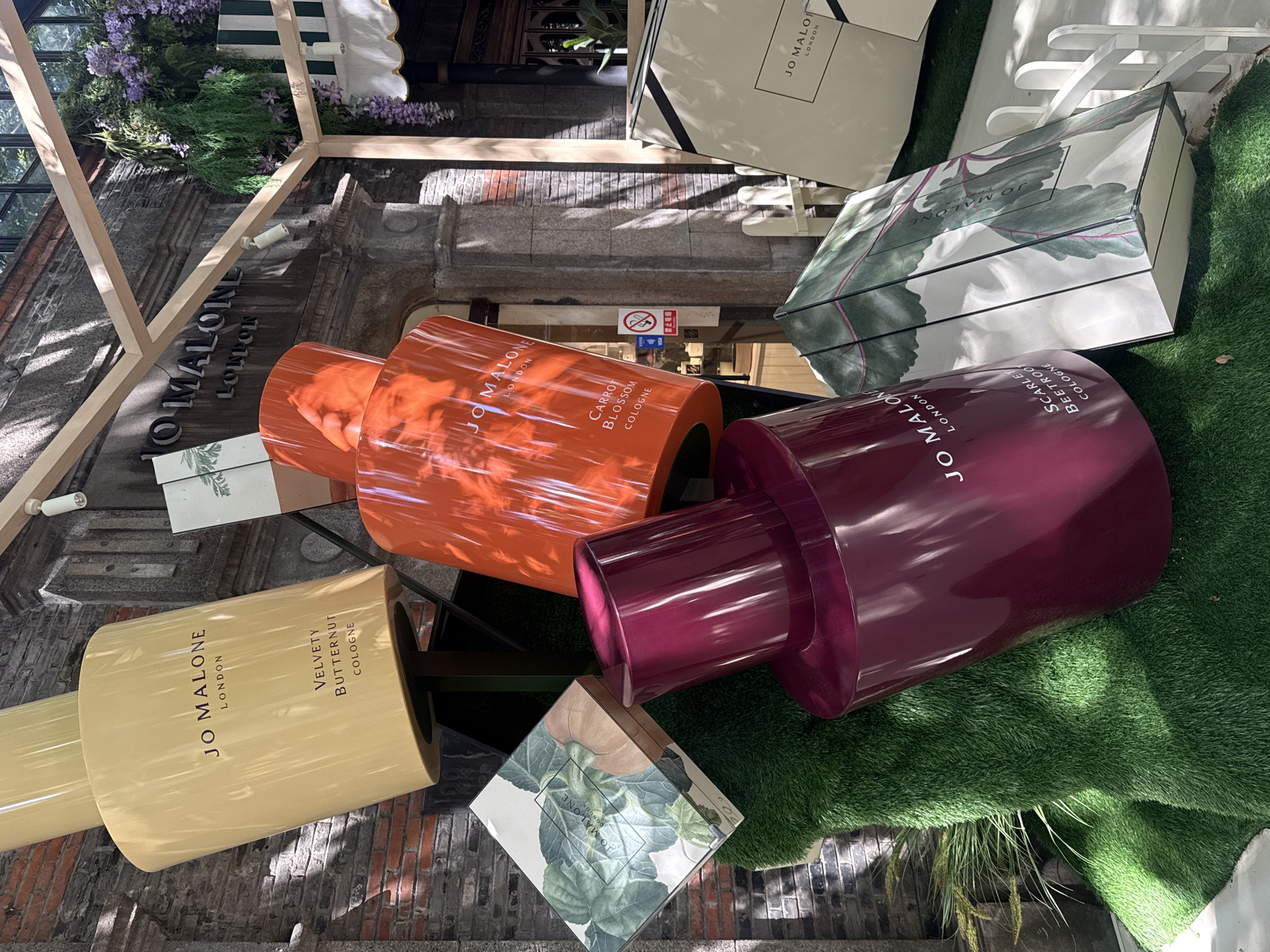 The image depicts a whimsical outdoor display of oversized Jo Malone London perfume bottles and packaging, set against the backdrop of a Jo Malone boutique in Shanghai, China. The display features several large, brightly colored perfume bottles, including a yellow one labeled "Velvety Butternut Cologne," an orange one that appears to say "Carrot Blossom Cologne," and a deep red one with "Scarlet Beetroot Cologne" visible. These are interspersed with large, square boxes designed to look like Jo Malone product packaging, adorned with botanical illustrations. The entire display is set on artificial grass, creating an immersive and playful atmosphere. In the background, the architecture of the Jo Malone store is visible, with its distinctive signage and a lush floral arrangement hanging from the building. The scene appears to be captured during daylight hours, with sunlight casting shadows on the display and the store facade. There are no people prominently featured in the foreground, suggesting the focus is solely on the product display. The setting implies a commercial or promotional event, possibly a store opening or a seasonal campaign, designed to attract attention and engage potential customers.