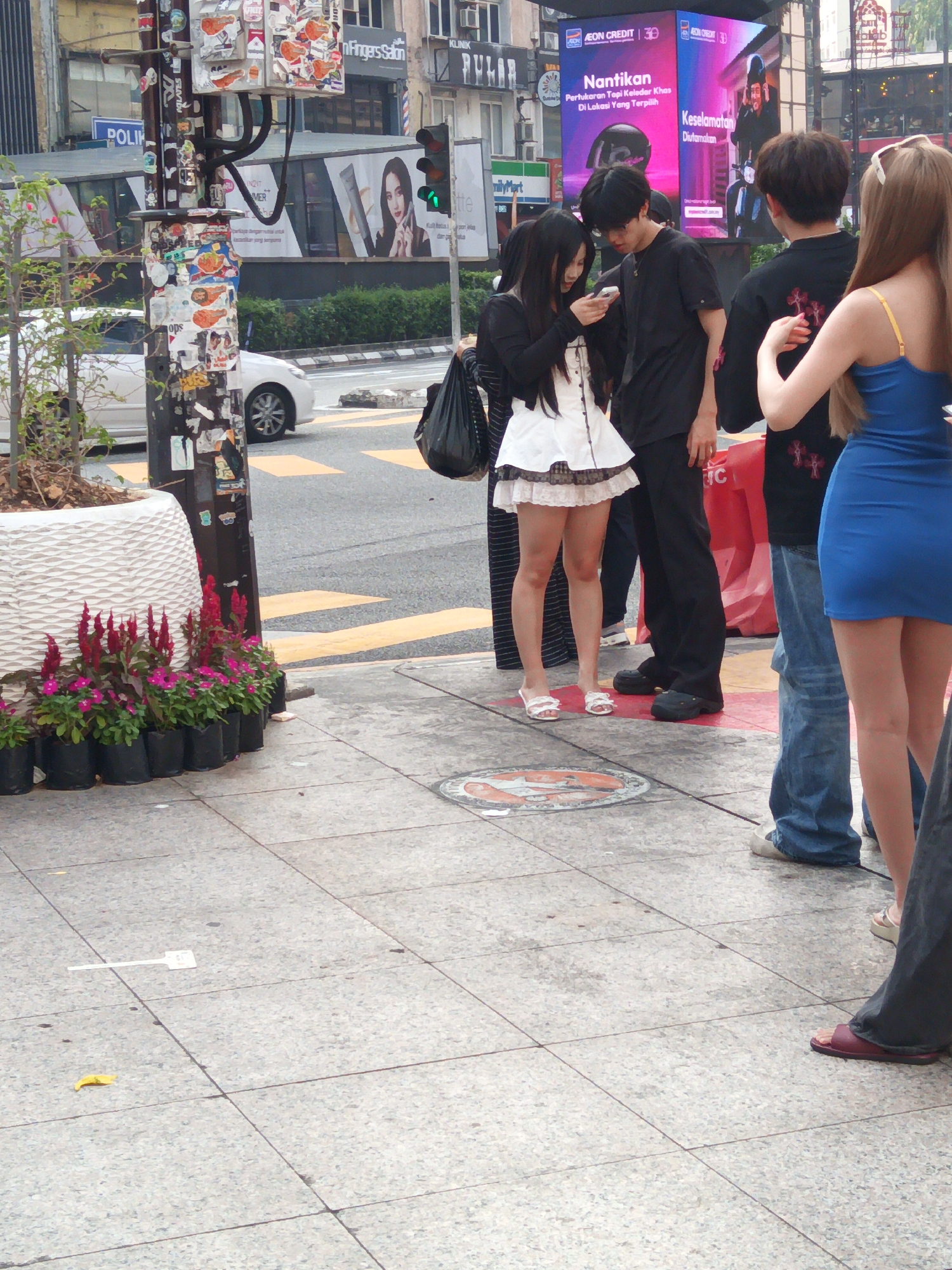 The image shows a street scene in Kuala Lumpur, Malaysia. A group of young adults are standing on a sidewalk next to a road with yellow crosswalk markings. In the foreground, a woman in a white and black dress with black hair is looking at her phone. To her left, a man in a black t-shirt and dark pants is also looking down. To the right of the woman, a man in a black shirt with red detailing on the back is wearing jeans, and a woman in a bright blue dress is visible from the waist down. The background features a traffic light, a utility pole covered in stickers and advertisements, and buildings. A white car is parked on the side of the road. Several potted plants are arranged along the sidewalk.
