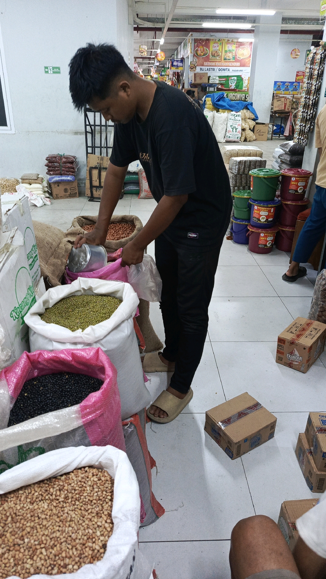 The image captures a scene inside a market or store in Ponorogo, Indonesia, likely a grocery or bulk food seller. The main subject is a young man wearing a black t-shirt and black pants, with tan slides on his feet. He is actively engaged in scooping what appears to be peanuts from a large burlap sack into a pink plastic bag. In front of him are several large sacks of dried goods: one filled with green mung beans, another with black beans, and a third with peanuts. Behind these, more sacks of food items are visible, along with stacks of smaller packages and various colored buckets, possibly for storing or selling grains and legumes. The environment appears to be an indoor retail space with tiled flooring and overhead lighting, common in Indonesian markets. The background shows shelves stocked with products and signage, further indicating a commercial setting. Another person's legs, wearing a light-colored shirt and shorts, are visible in the lower right of the frame, suggesting another customer or vendor. The overall impression is a busy, functional marketplace environment focused on the sale of bulk dry goods.