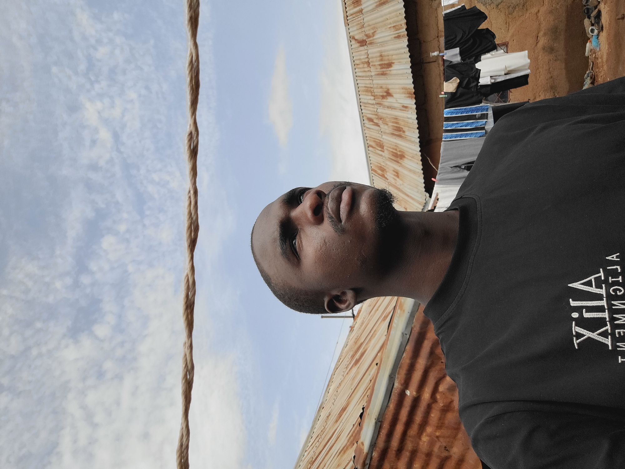 A low-angle shot in Bukuru, Nigeria, reveals a Black male wearing a black T-shirt with white lettering ("XHA ALIGNMENT"). He is looking upwards, against a backdrop of a partly cloudy sky. A thick, brown rope is positioned to the left. Behind him, a corrugated metal roof shows signs of rust. Clothes are hanging on a line in the background. The ground appears to be made of red earth.