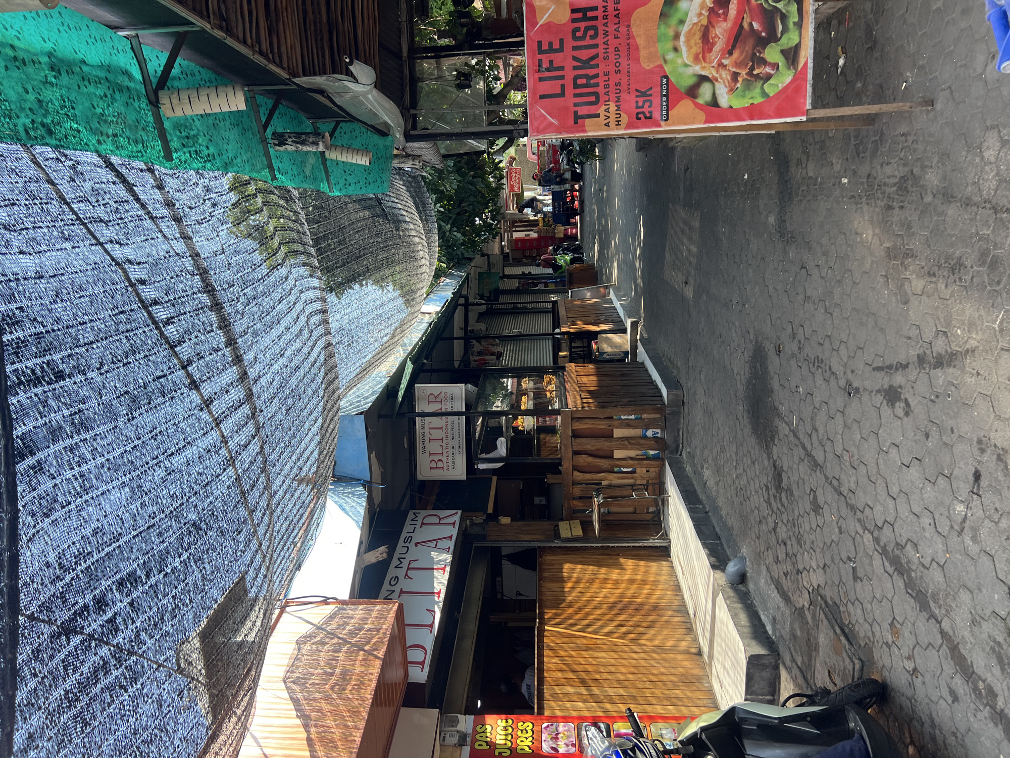 The image depicts a street scene in Kuta, Indonesia, likely a market or a row of food stalls. A shaded walkway, covered by a dark mesh canopy, runs through the scene. On the left, a food stall with wooden facade displays "BLITAR" signage. To the right, another stall is visible, also featuring food displays. A red sign promoting "LIFE TURKISH" food is prominent in the foreground, advertising shawarma, hummus, soup, and falafel, with a price of "25K" and an "ORDER NOW" call to action. A motorcycle is parked on the left side of the walkway. The ground is paved with interlocking hexagonal stones, some of which appear wet. Trees and foliage are visible in the background, suggesting an outdoor environment. The overall impression is that of a bustling commercial area, likely catering to tourists and locals.
