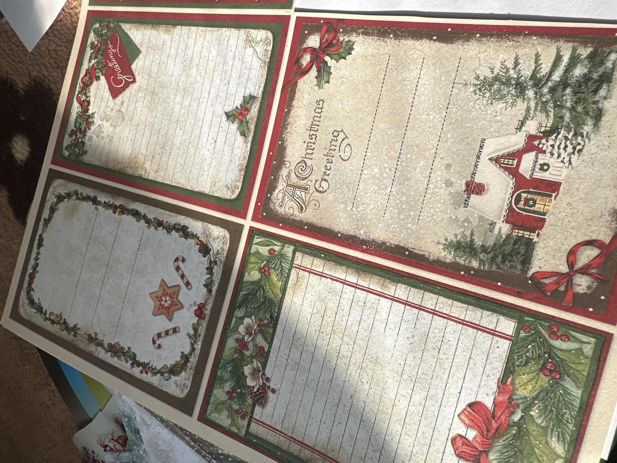The image shows a sheet of decorative paper with four different Christmas-themed designs. Each design is a bordered rectangle with lined space for writing. The upper-left design includes a small red tag with the word "Greetings" written on it. The upper-right design has the phrase "A Christmas Greeting," along with an image of a house and pine trees. The lower-left design includes a star and candy canes. The lower-right design has flowers and pine tree leaves on it.