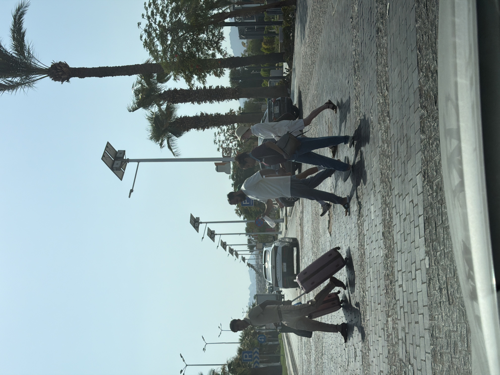 In Sharm El Sheikh, Egypt, on a clear, sunny day, a group of four pedestrians, two men and two women, cross a paved road at a designated crosswalk. The man on the far left is wearing a light-colored shirt and pants and is pulling a red suitcase. Beside him, a woman in a dark shirt and jeans is walking with a shoulder bag. Next to her, a woman in a white dress and a straw hat is walking. The man on the far right is wearing a light-colored shirt and dark pants, holding a white bag. Behind them, a white sedan is stopped at the crosswalk, with other vehicles and palm trees visible in the background. Streetlights with solar panels are lined up along the road. The ground is paved with grey interlocking bricks.