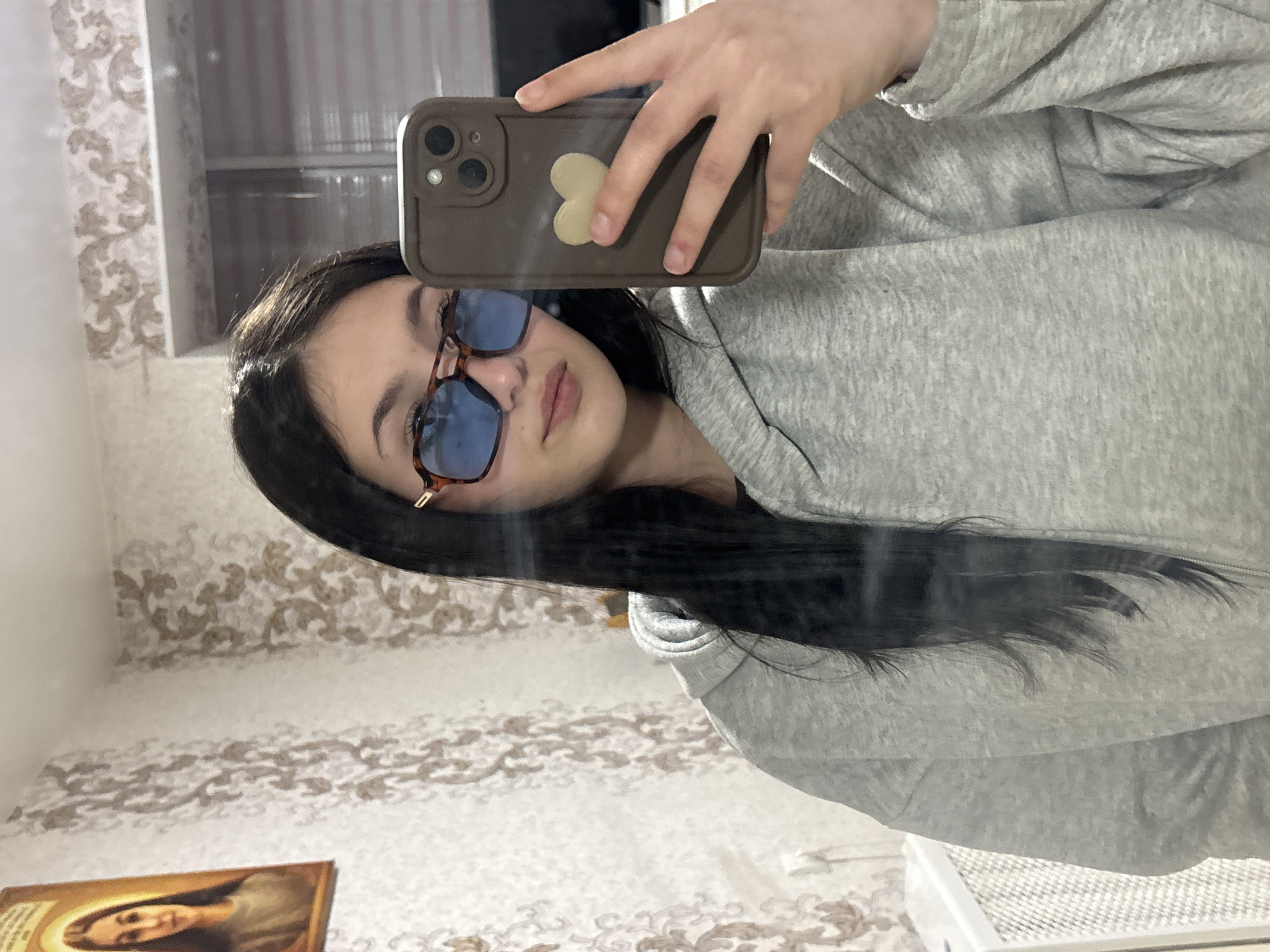 A woman is taking a selfie in a mirror. She is wearing a gray hoodie and blue-tinted sunglasses with brown frames. Her phone has a brown case with a cream-colored heart on the back. The background includes a patterned wall and a shelf with various items, including what appears to be an icon. The location Фоки, Russia is not discernible from the image.
