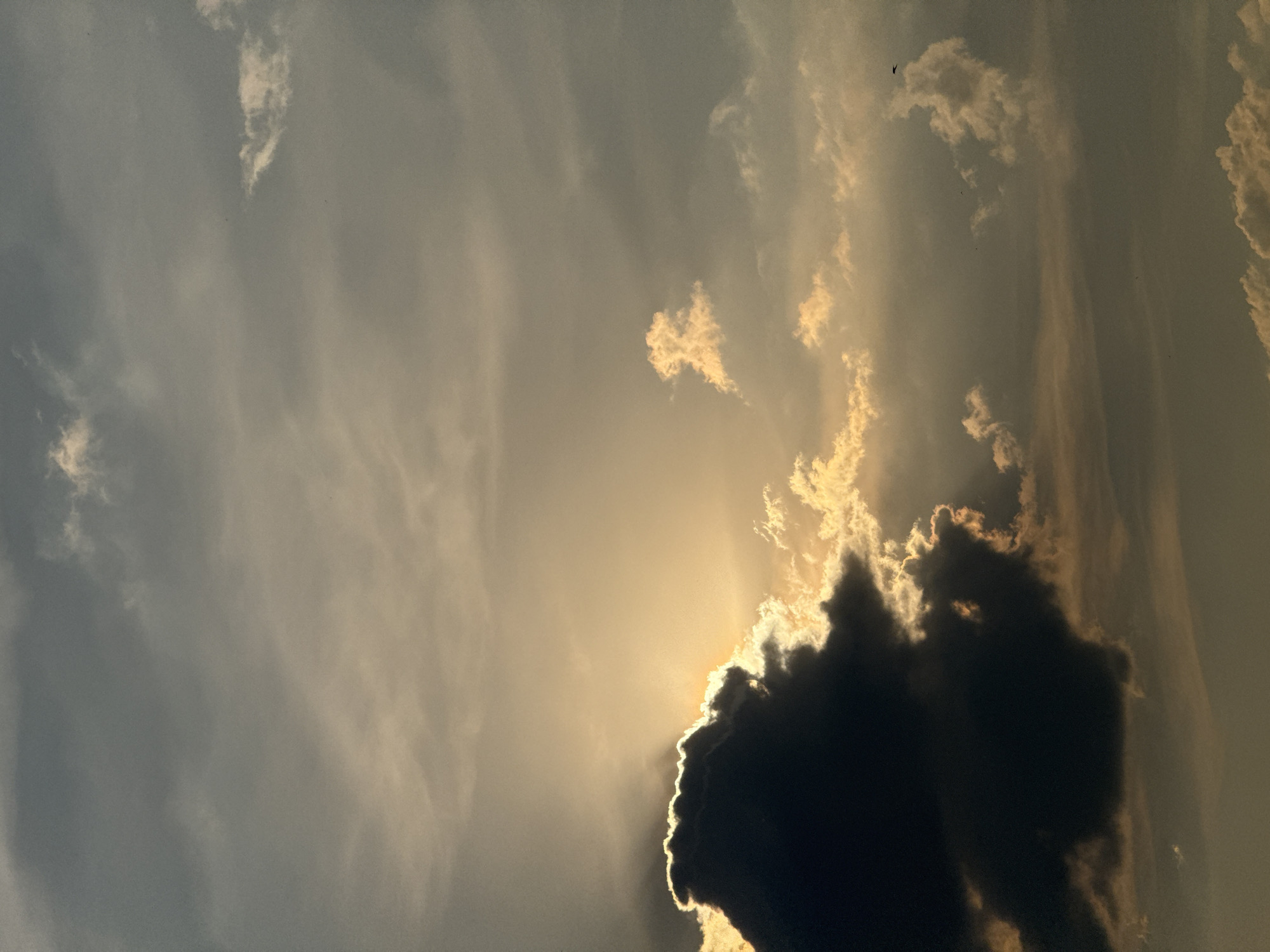 The image shows a dramatic sky with a large, dark cloud in the lower left corner, partially silhouetted against the setting sun. Rays of sunlight are breaking through the edges of this cloud and illuminating other smaller clouds in the sky with a golden hue. The overall sky is a mix of grey and golden tones, suggesting either sunrise or sunset. There are no identifiable landmarks or urban features, therefore, the location in Moscow, Russia cannot be determined from the image. There are no people, subjects, or objects depicted in the image other than clouds.