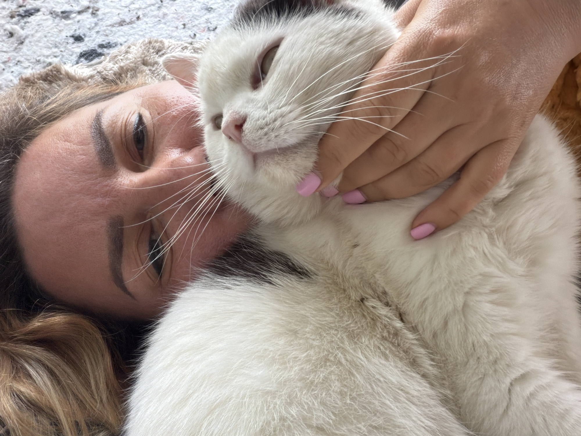 A woman lies on a surface, possibly a carpet or blanket, next to a white cat with some black markings on its head and back. Her right hand is on the cat’s face. The woman has brown hair, dark eyebrows, and one eye visible. The cat has closed eyes. The setting is not discernible as being in Leningradskaya, Russia.