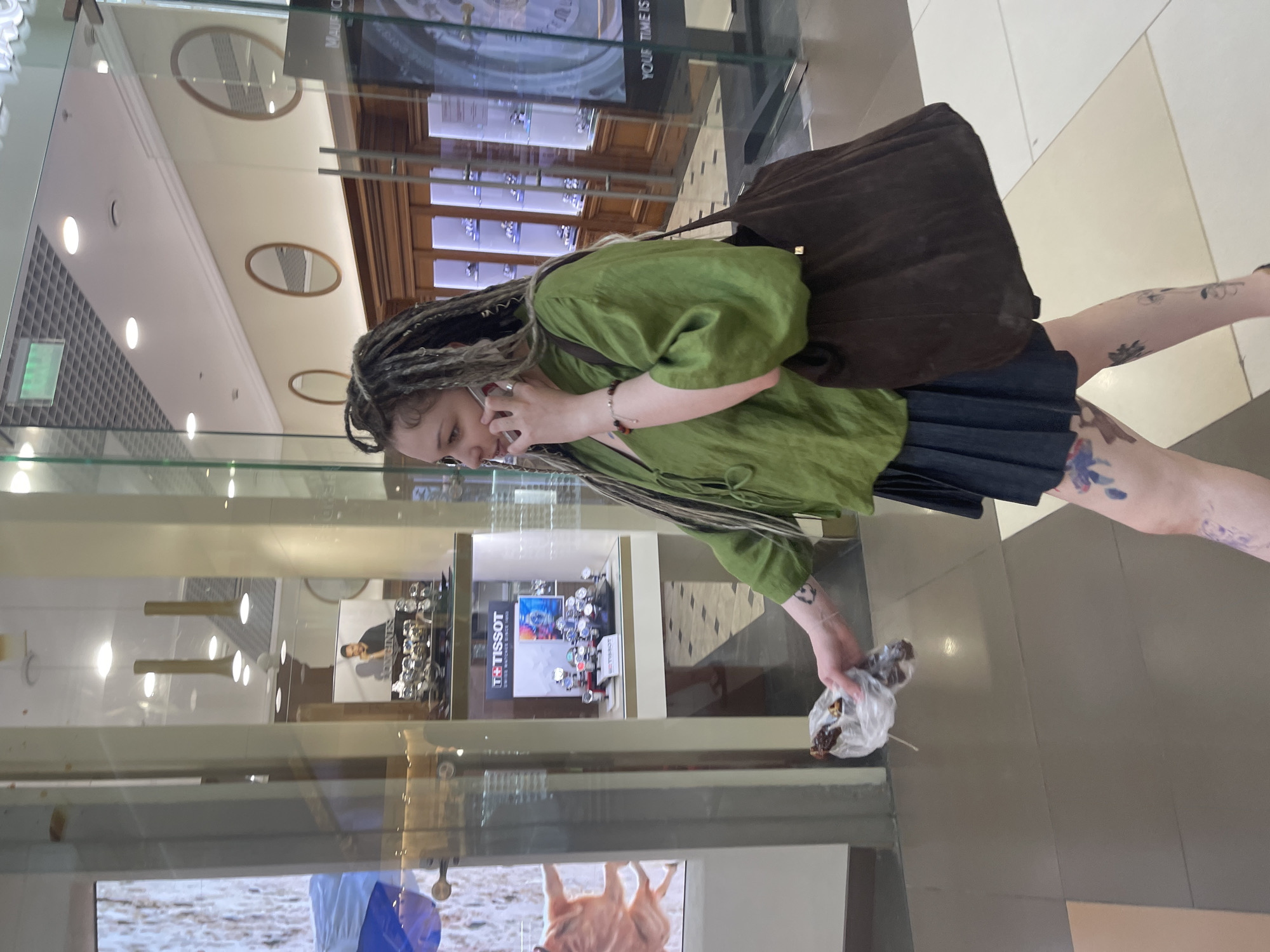 A young woman with dreadlocks, wearing a green blouse and a dark pleated skirt, walks through a shopping mall while talking on her phone and holding a plastic bag containing food. She has tattoos on her legs. The background shows a jewelry store with display cases of watches. The floor is tiled in a checkered pattern. The lighting suggests it is daytime indoors. The scene appears to be in a shopping mall in Moscow, Russia.