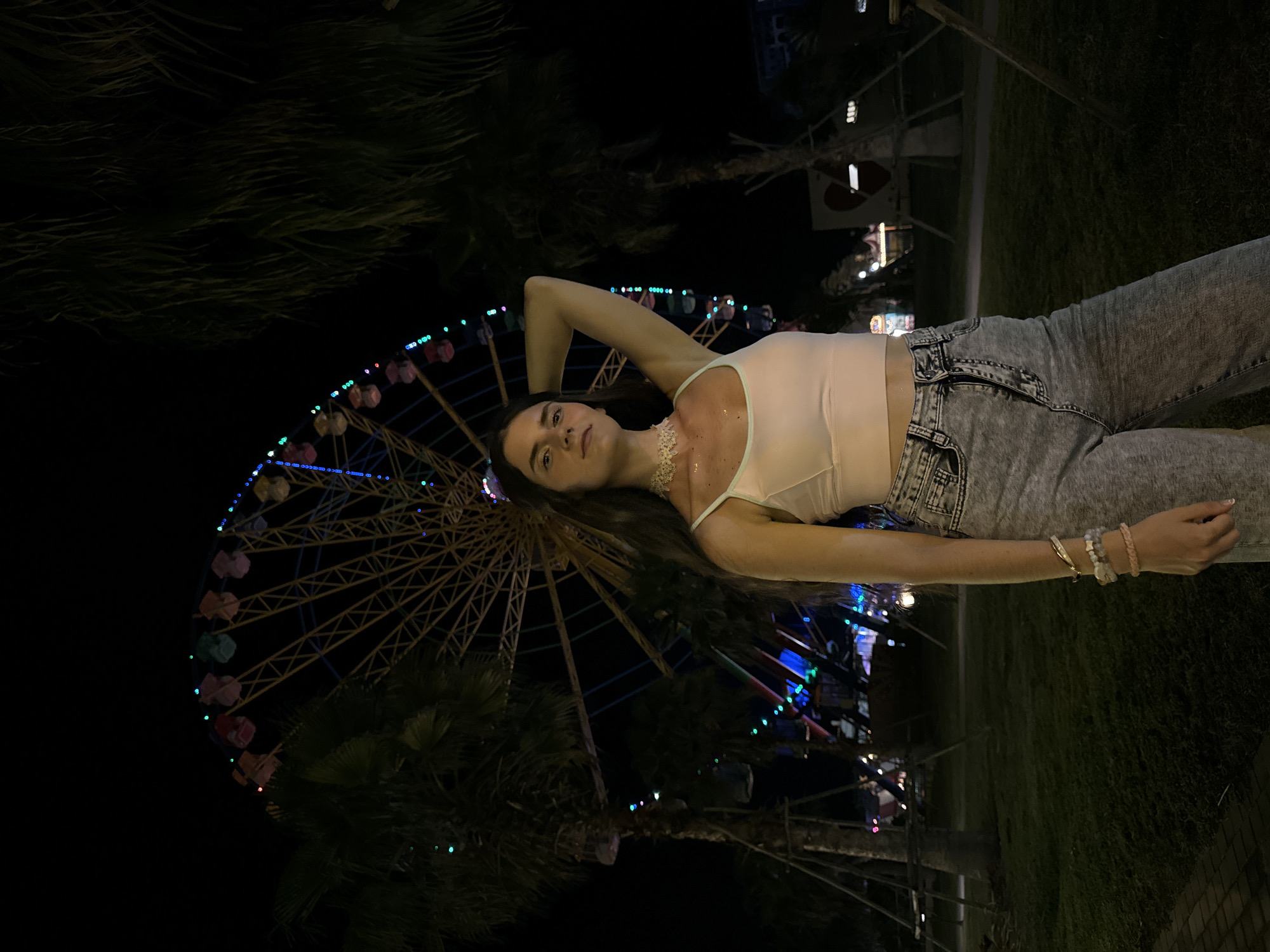 A young woman stands in front of a Ferris wheel at night. She is wearing a light pink tank top, grey denim jeans, and multiple bracelets on her left wrist. Her right arm is raised behind her head. The Ferris wheel is brightly lit with colorful lights and is in the background, partially obscured by palm trees. The scene is set in a park or amusement area, with grass and trees visible in the foreground and background. The overall atmosphere is festive and urban, with the Ferris wheel suggesting a location like an amusement park or a nighttime fair. The image appears to have been taken at an outdoor venue in Tien Thanh Ward, Vietnam, indicated by the presence of tropical palm trees and potentially local amusement park elements.