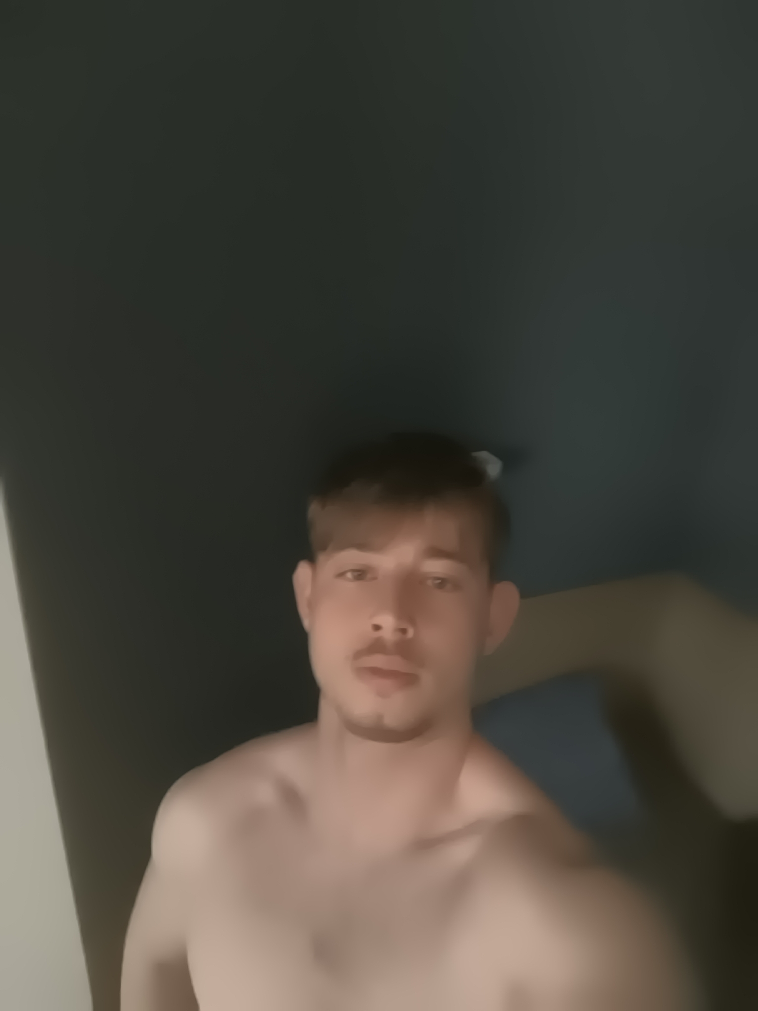 The media features a young man, appearing shirtless or topless, captured from the chest up. He has short, light brown hair, a light mustache, and a small goatee. His skin is fair, and he looks directly into the camera with a neutral expression.
The setting is an indoor space, likely a room, characterized by dim lighting. The background is mostly dark, appearing as a solid dark grey or dark teal surface, suggesting a wall or ceiling. A lighter, possibly white or light grey, vertical strip is visible along the left edge of the frame, which could be a doorframe or corner. To the right, behind the man's shoulder, a blurred blue or grey shape is faintly discernible, possibly part of furniture like a bed or pillow.
The image is somewhat blurry and grainy, which could be due to low light conditions or being a still frame from a video. No specific activity beyond posing for the camera is evident. The time of day appears to be evening or night given the subdued lighting. There is no visible text in the image. The location context provided for this media is Sofia, Bulgaria, although no specific visual cues within the image confirm this geographical detail.