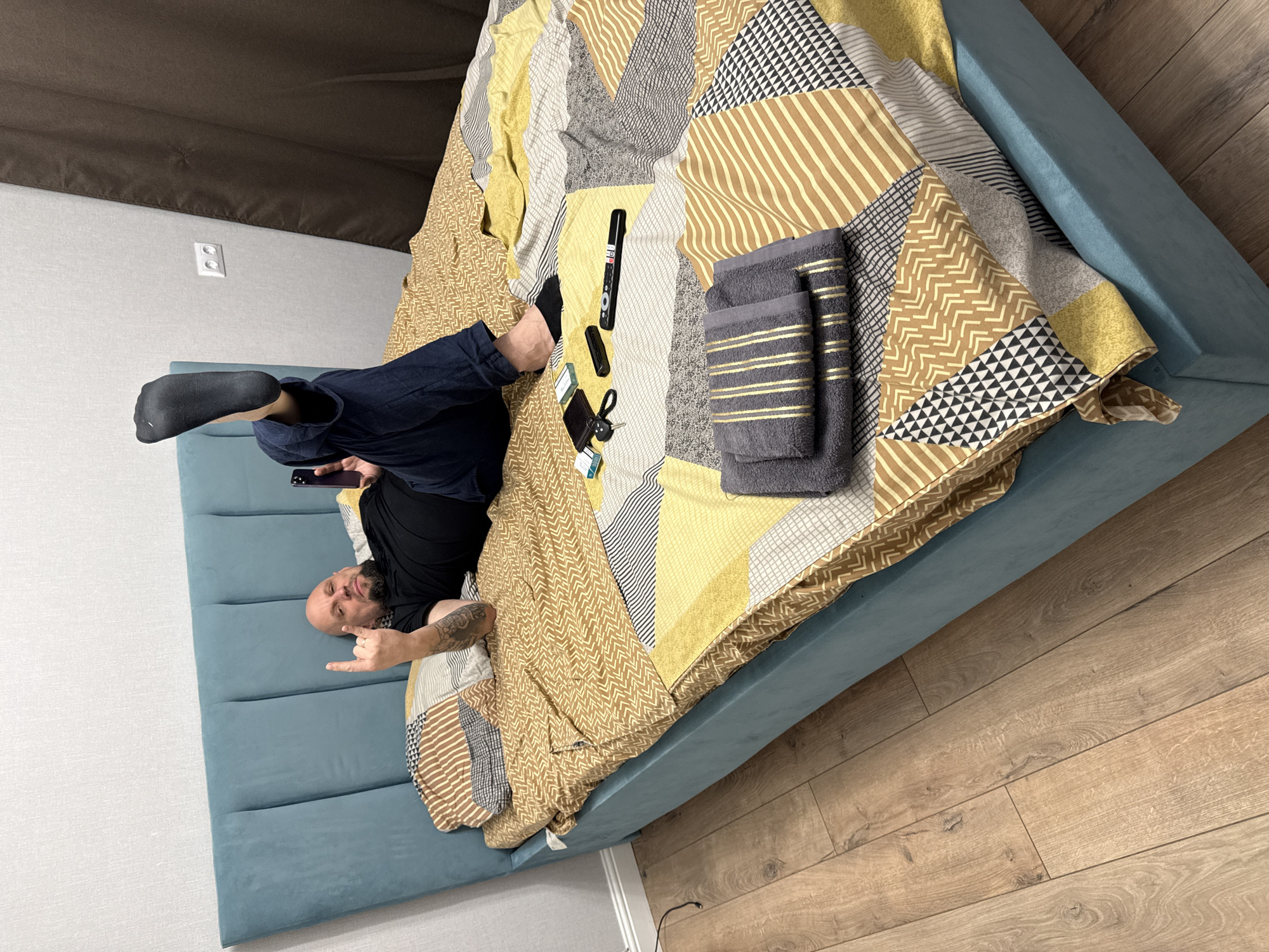A man is lying on a bed with a blue headboard and a patterned yellow, gray, and white comforter in a room with wood flooring and light gray walls. He is wearing a black t-shirt, dark blue jeans, and a black sock on his raised left foot. He is holding a black smartphone and making the "hang loose" sign with his right hand. On the bed next to him are a black TV remote, a black car key, a brown leather wallet, two boxes of cigarettes, and a stack of two gray towels with gold stripes. A white electrical outlet is visible on the wall behind the headboard.
The location cannot be determined from this image; there is no evidence to confirm it is in Ovoshchnoy, Russia.