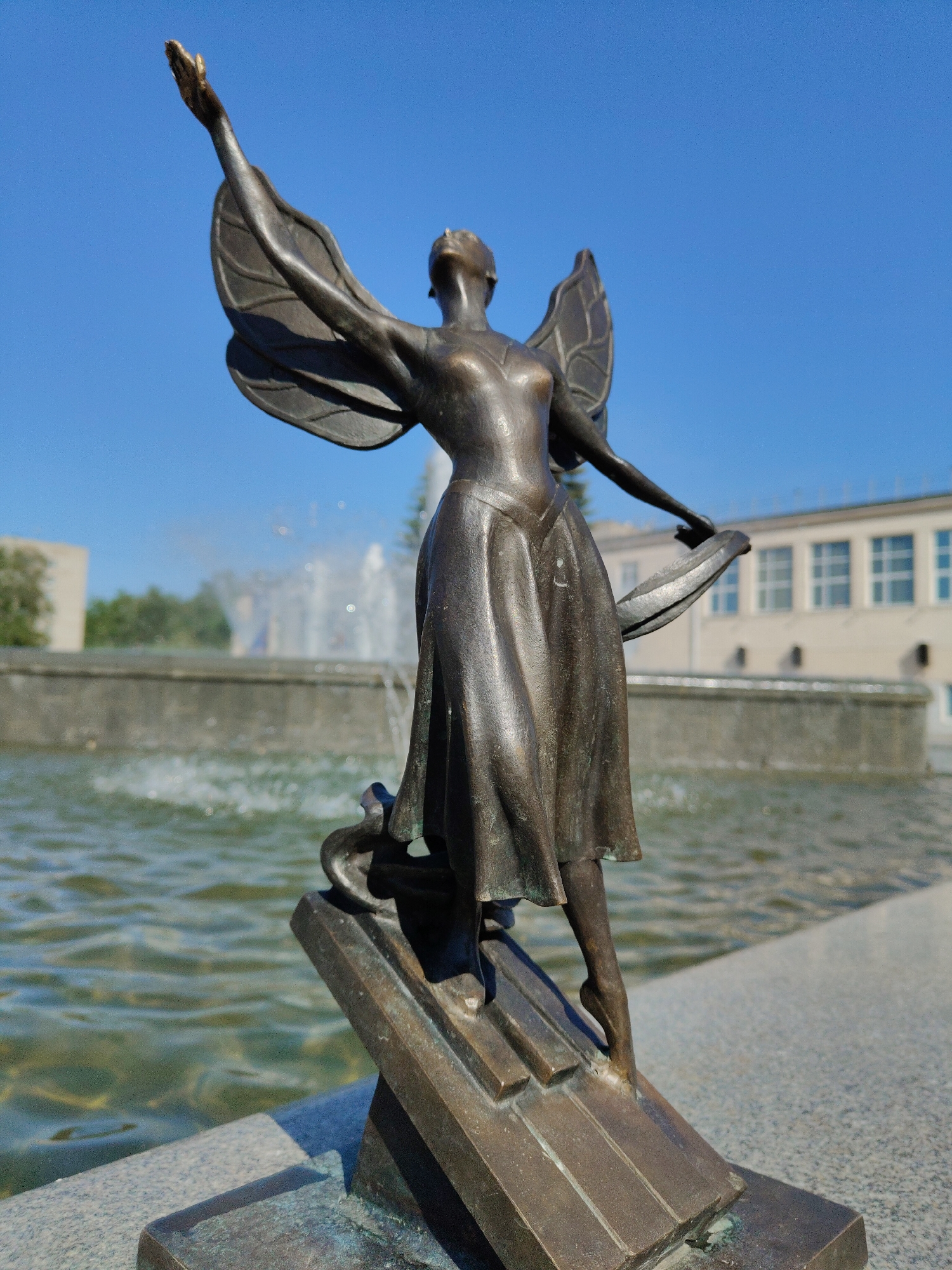 A bronze statue of a winged female figure stands in front of a fountain on a sunny day in an urban setting. The statue, likely commemorating a ballerina or dancer, has her arms extended, one reaching upwards, and a wing extending from each shoulder. She is clothed in a long dress and appears to be in motion.
The statue is mounted on a bronze base that resembles stylized piano keys or steps. In the background, a fountain is actively spraying water. Behind the fountain, blurred buildings and trees suggest a city or park landscape. The statue is placed on a concrete ledge or platform, and the background is a clear blue sky.